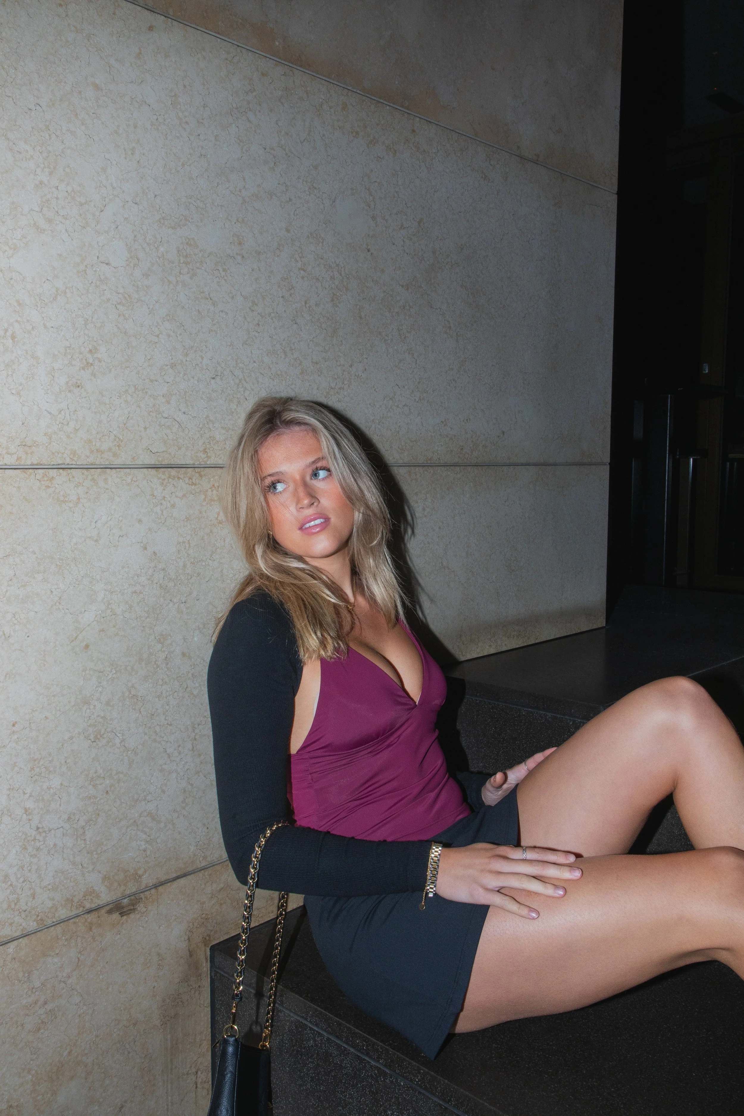 Young woman with blonde hair sitting on a black step, leaning against a beige wall, looking to her right, wearing a burgundy top, black shorts, and a black cardigan with a chain-strap purse hanging from her shoulder.