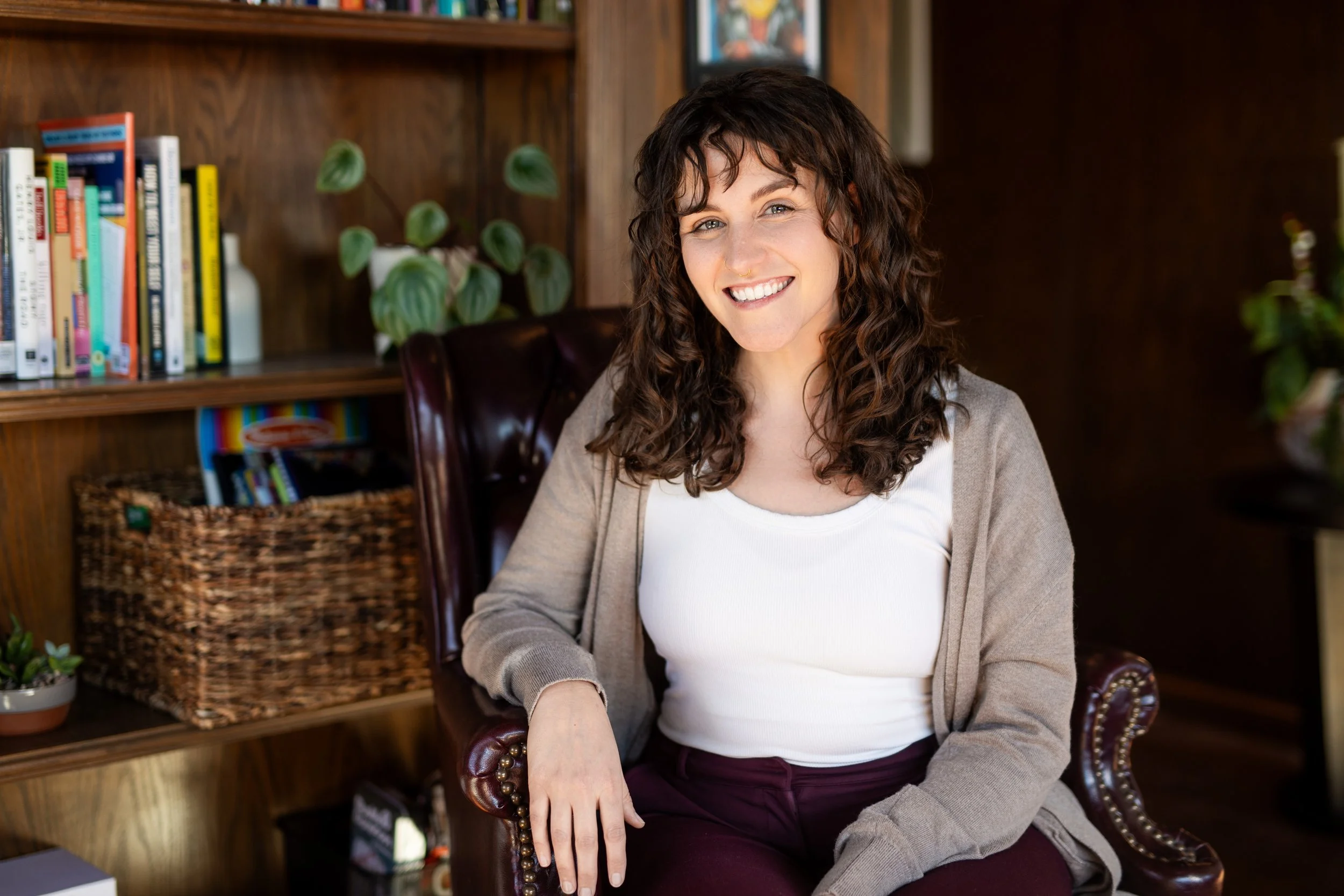 A woman smiling looks forward with brown hair a white t shirt and a tan cardigan.