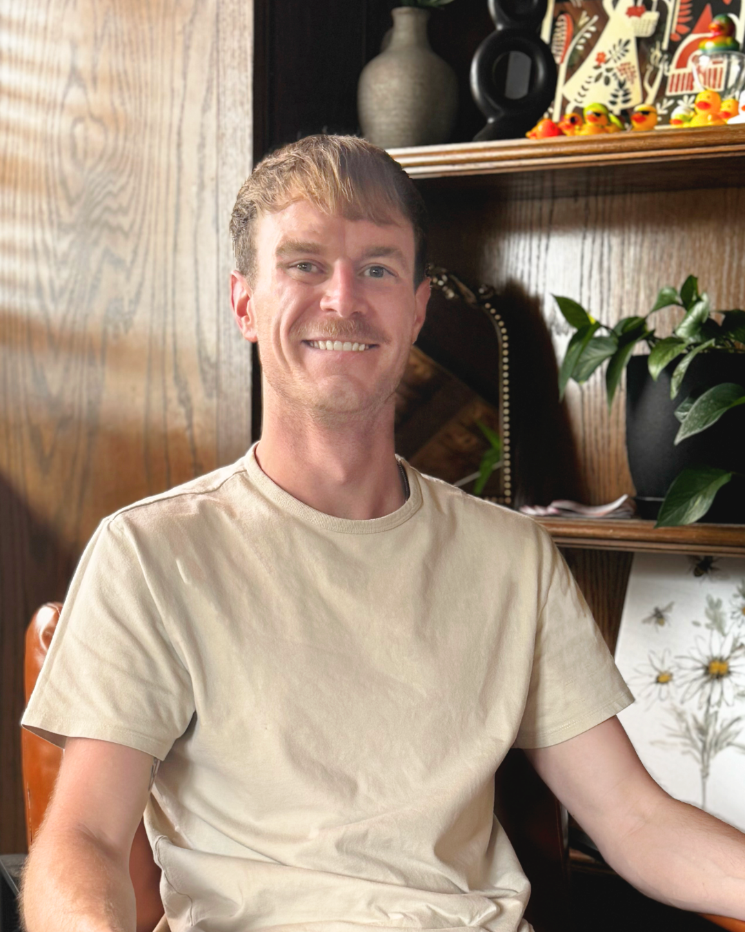 A man looks forward sitting in front of a bookshelf smiling.