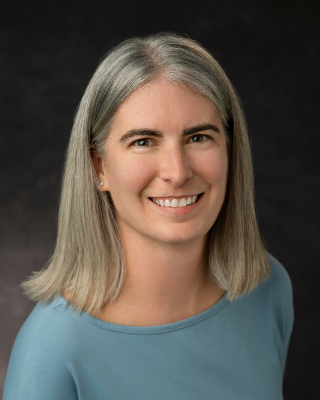 a woman with light blonde/gray hair looks forward in a blue top smiling.
