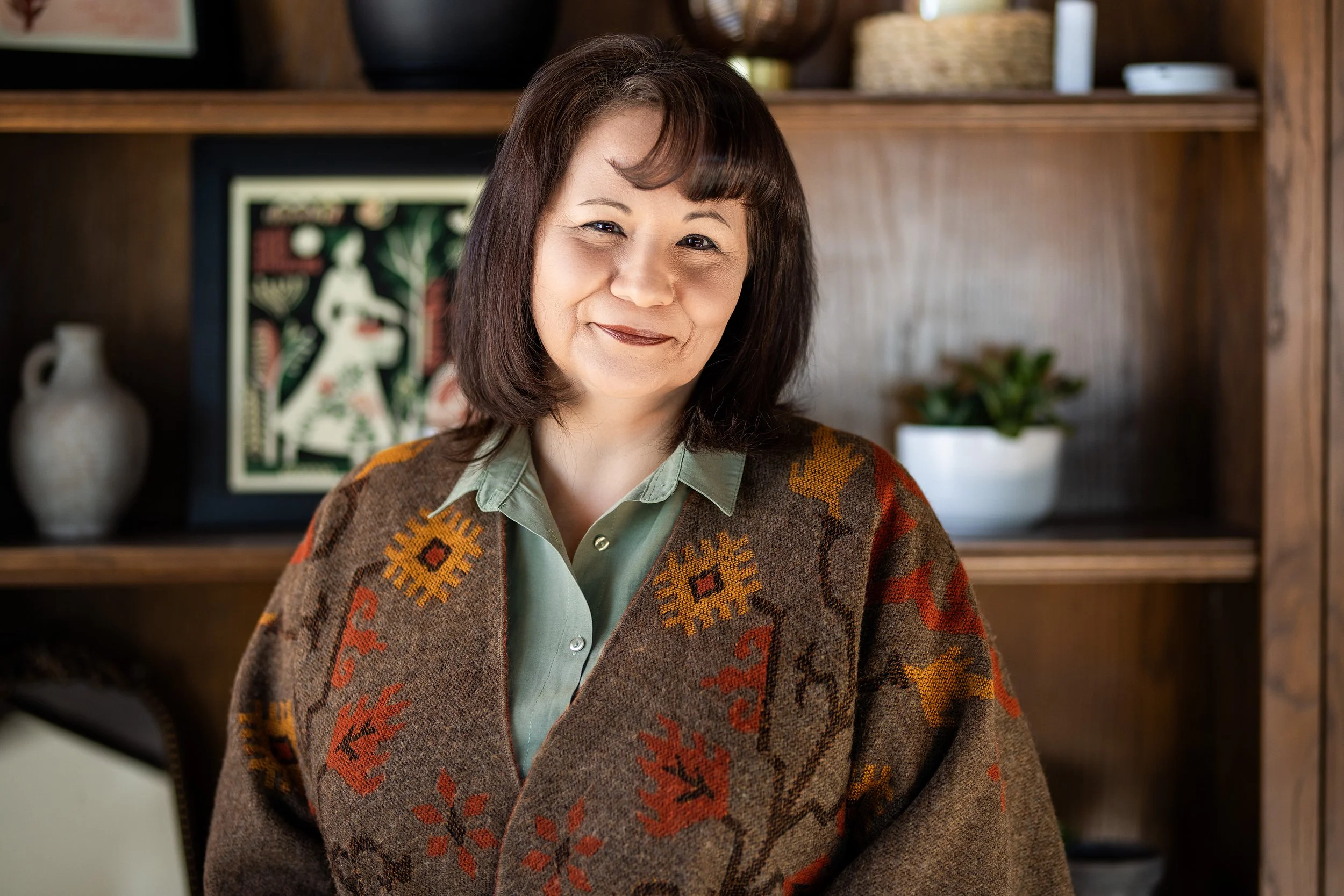 Young woman counselor/therapist with shoulder-length light brown hair, smiling, wearing a white blouse with red embroidery on the chest, standing outdoors.