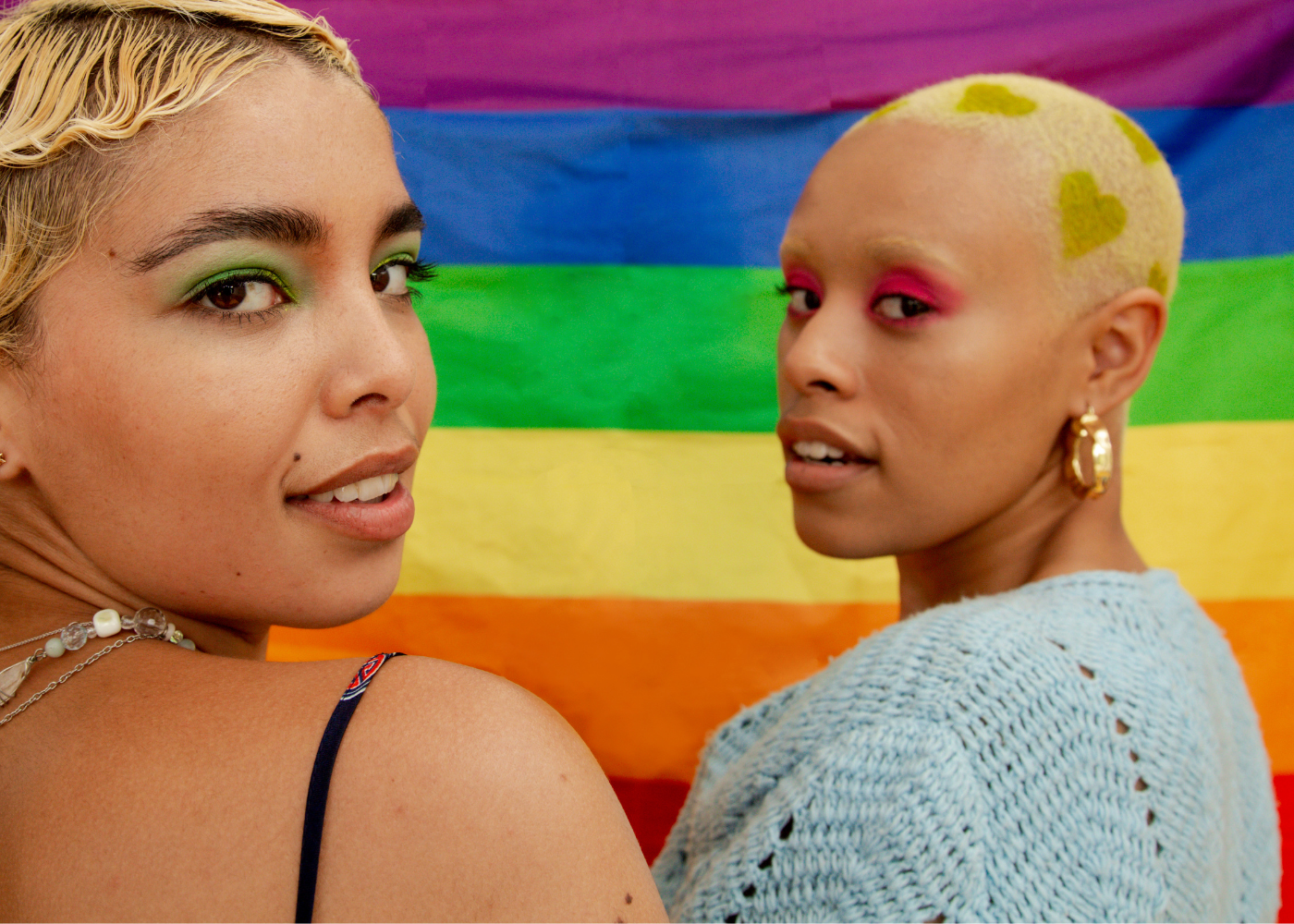two brown femme queer lgbtq folks look over their shoulder at the camera with a rainbow flag in the background. photo by Shann Daniels