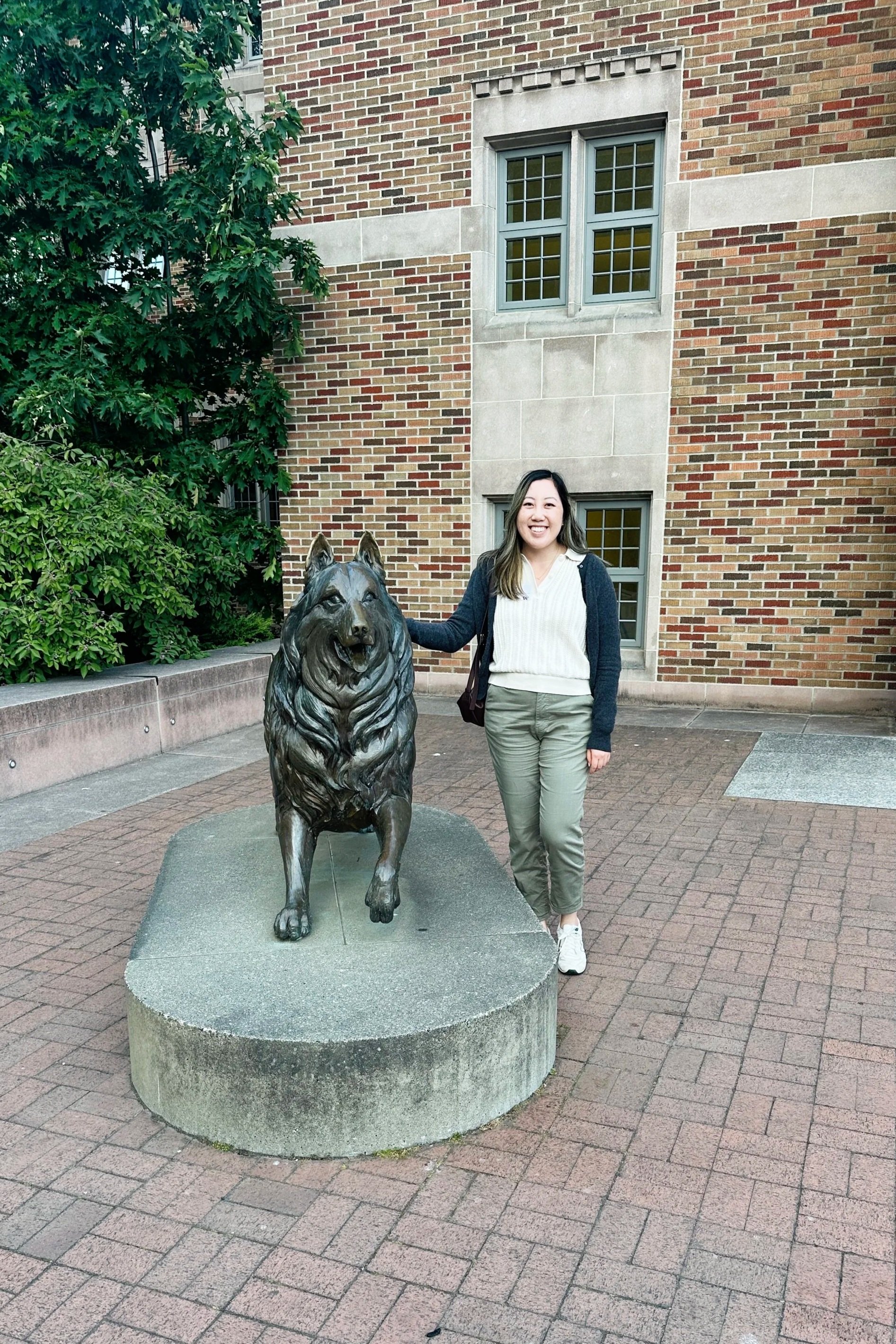 A woman standing next to a bronze statue of a dog on a stone pedestal outside a brick building with green trees in the background.