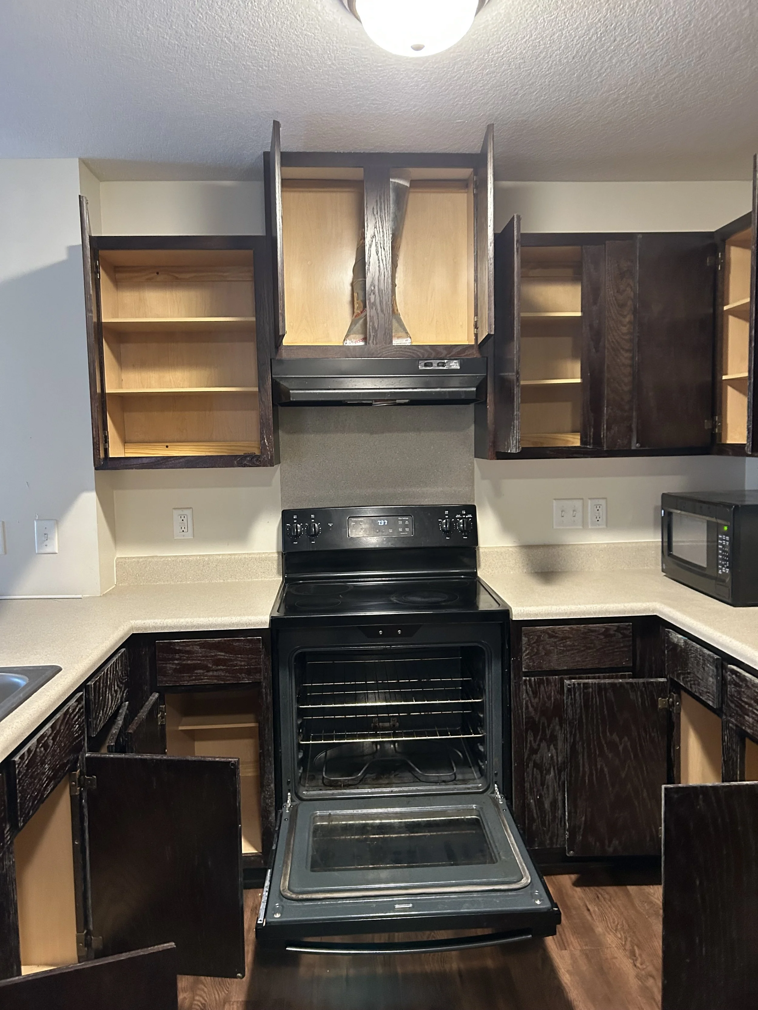 Open kitchen cabinets surrounding a black electric stove with an open oven door, a microwave on the right counter, and an empty open cabinet below it. The cabinets are a mix of dark and light wood finish.