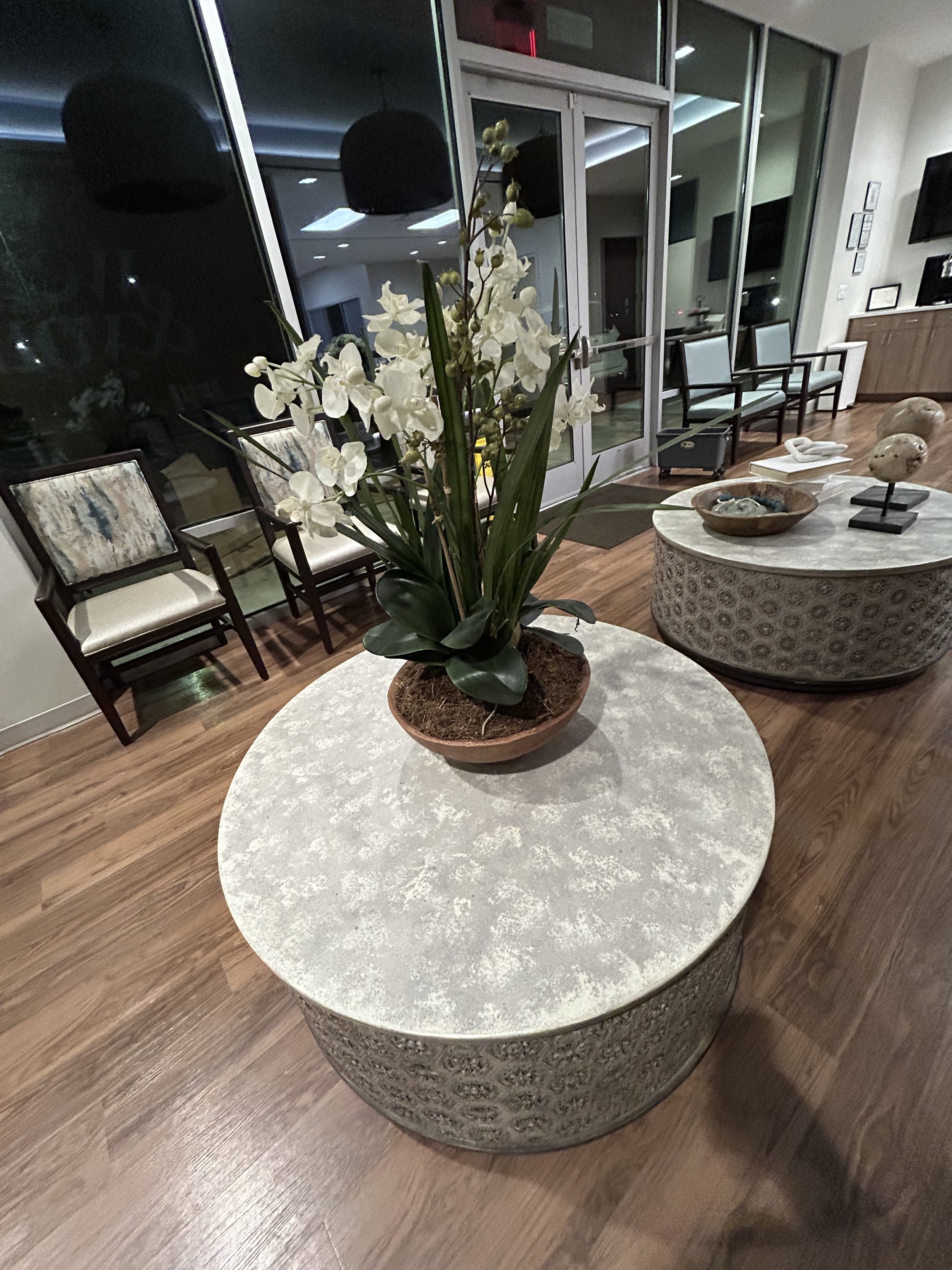 Living room with a round marble-top table with a potted white orchid plant, wooden floors, and additional decorative tables with sculptures, while chairs and windows are visible in the background.