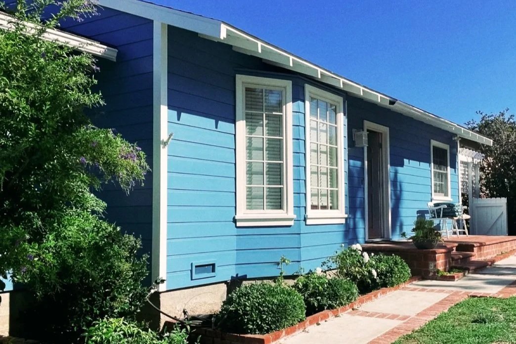 A blue house with white trim and multiple windows, a brick steps leading to a door, surrounded by green bushes and a small patio area.