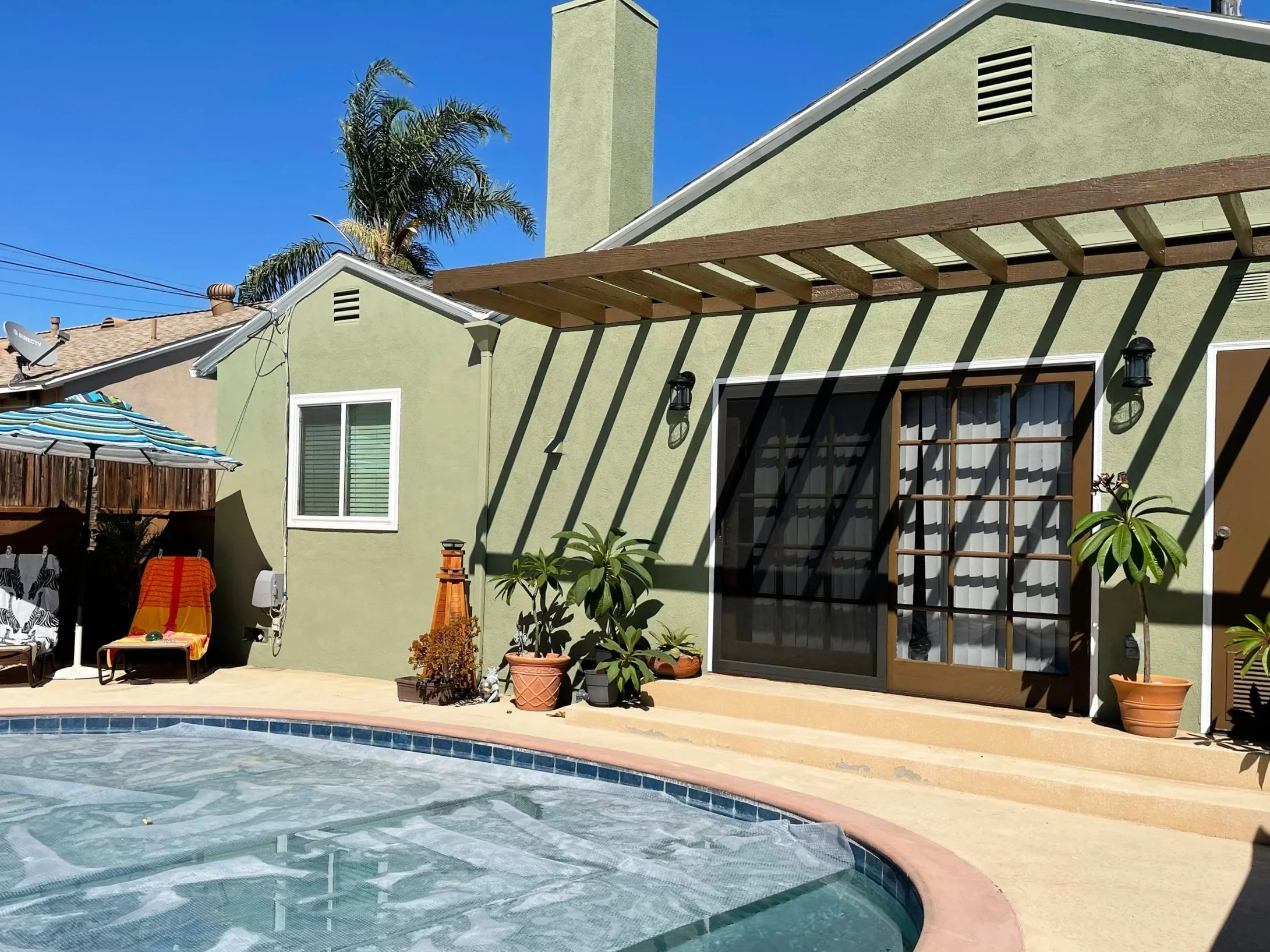 Backyard scene with a swimming pool, patio furniture including a striped umbrella and lounge chairs, and a two-story house painted light green with potted plants and a wooden pergola casting shadows.