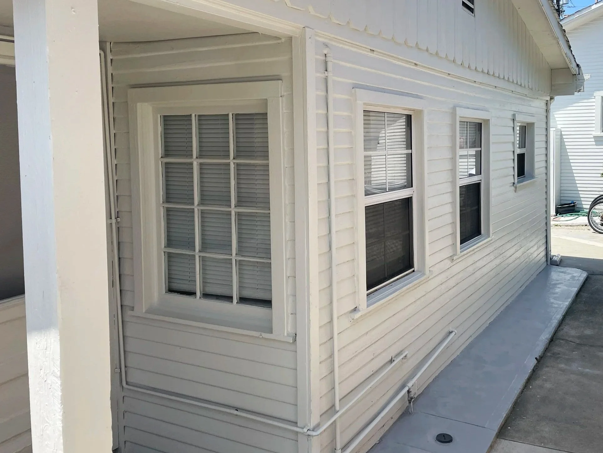 Exterior view of a white house with multiple double-hung windows with blinds. The house has horizontal siding and a small porch area with white pillars, concrete walkway, and some garden hoses on the side.