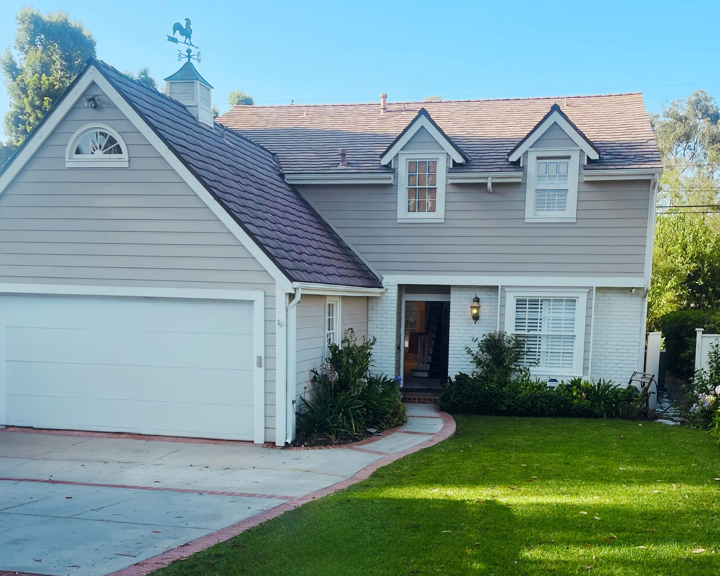 Front view of a two-story house with a garage, landscaped yard, and clear blue sky, featuring white siding, multiple windows, and a small porch with plants.