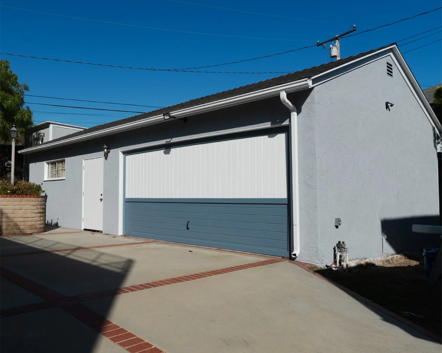 A white garage with a blue and white sectional door, attached to a gray house with a small window and door, on a concrete driveway with brick accents.