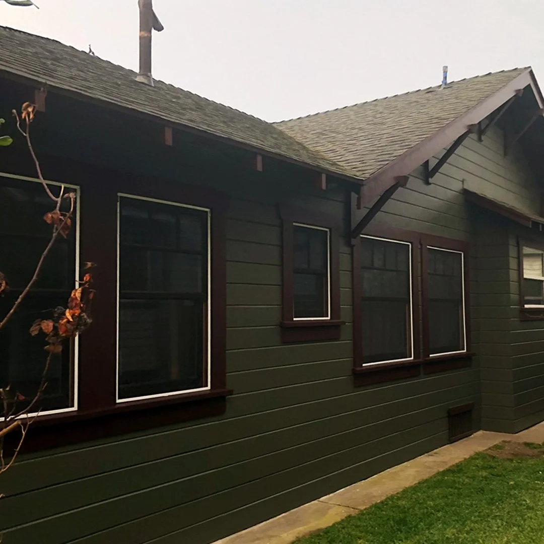 Backside of a house with green siding and four windows with brown trim, overcast sky, grass and sidewalk in foreground.