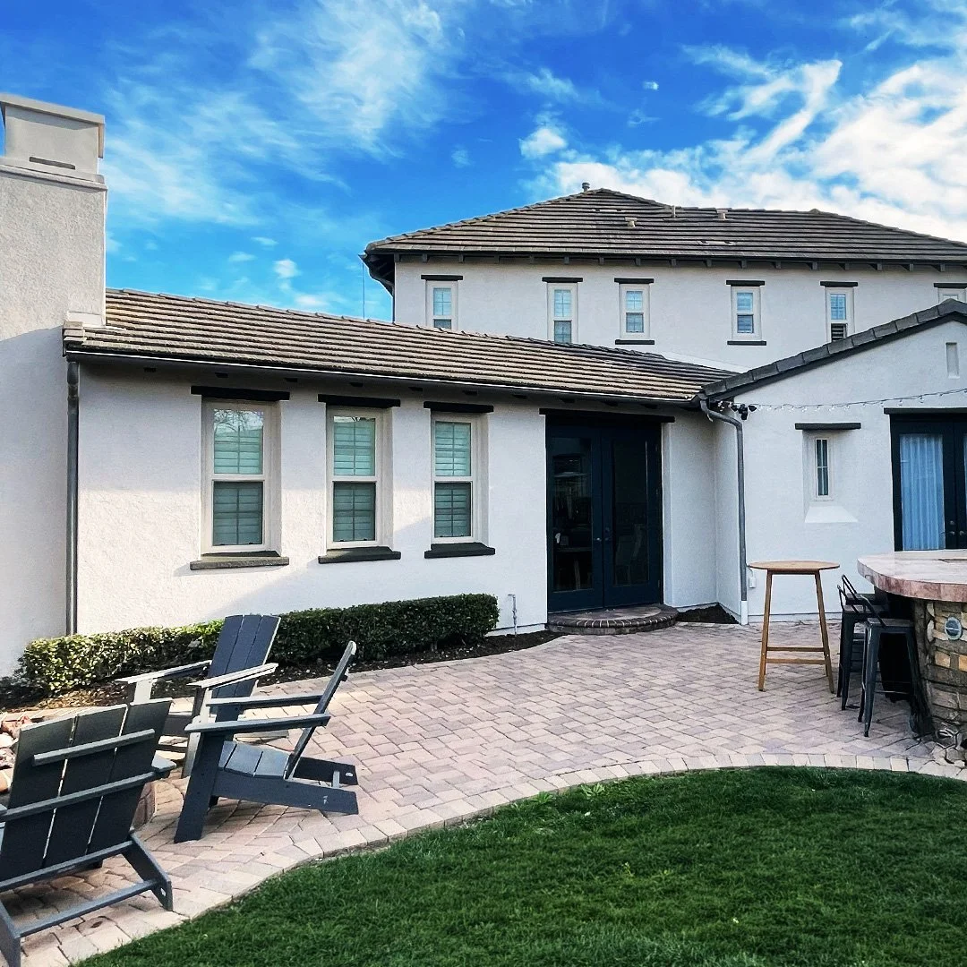 A backyard patio with black chairs, a small table, and a low brick wall, attached to a white house with multiple windows under a blue sky with some clouds.