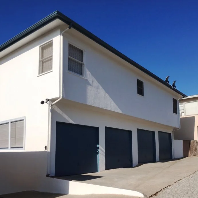 A white two-story house with a driveway, blue garage doors, and small windows, against a clear blue sky.