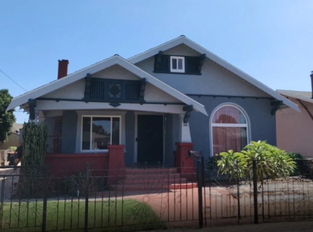 A blue house with a front porch, red accents on the porch columns, a large arched window with curtains, a small dormer window on the roof, and a front yard with a green shrub, a small tree, and a black metal fence.