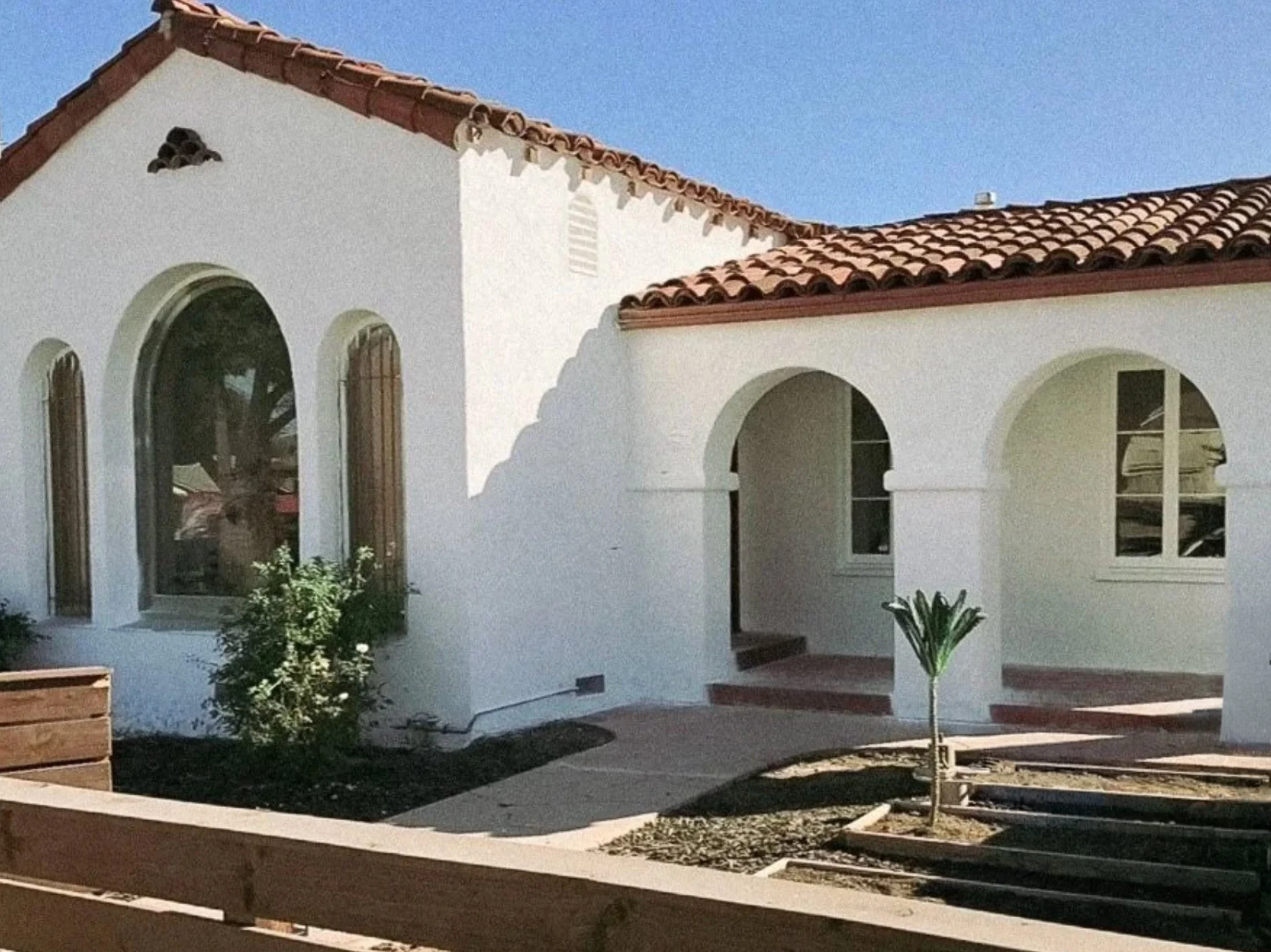 White house with red tile roof, arched windows, and a small tree in the front yard during daytime.