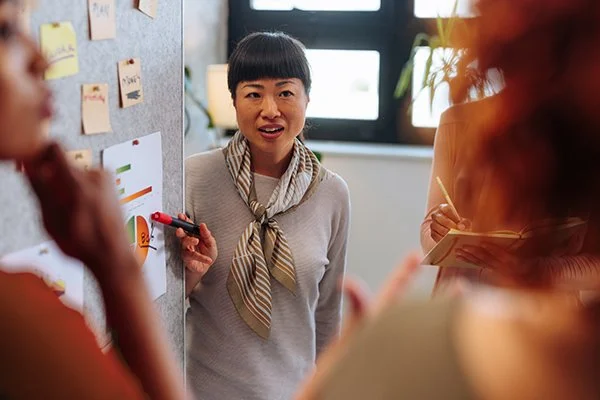 Woman with short black hair and striped scarf speaking in a meeting room with sticky notes on the wall.