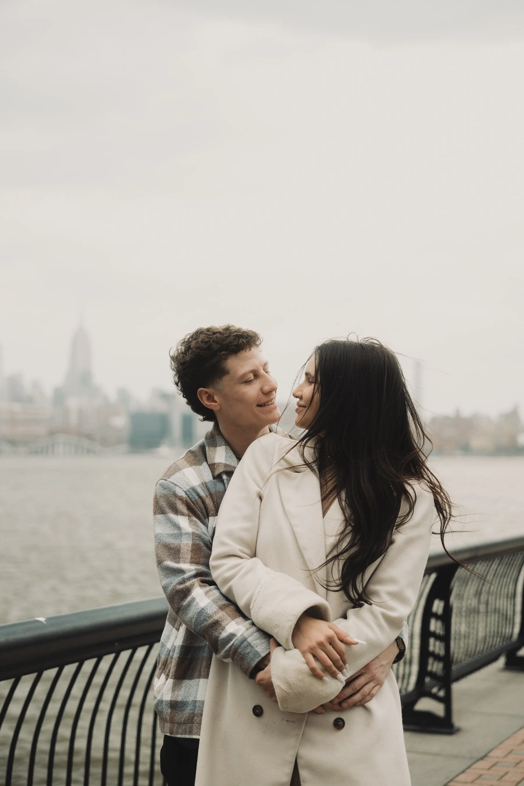 A young couple embraces by the waterfront with a city skyline in the background, on an overcast day.