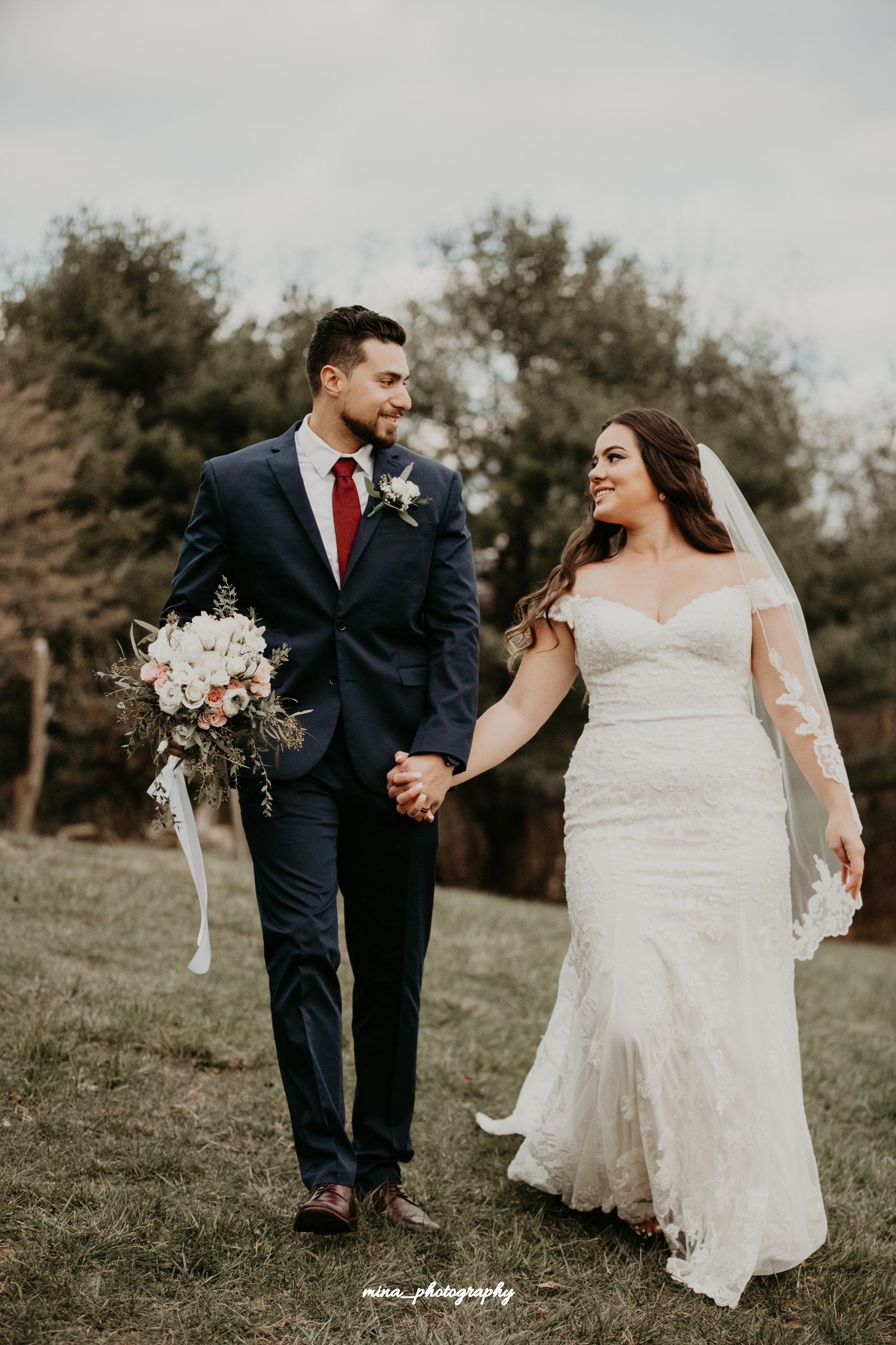 A bride and groom walking hand in hand outdoors on a grassy area during their wedding day, with tall trees in the background. The bride is wearing a white lace wedding gown and veil, and the groom is dressed in a navy suit with a white shirt and red tie, holding a bouquet of flowers.