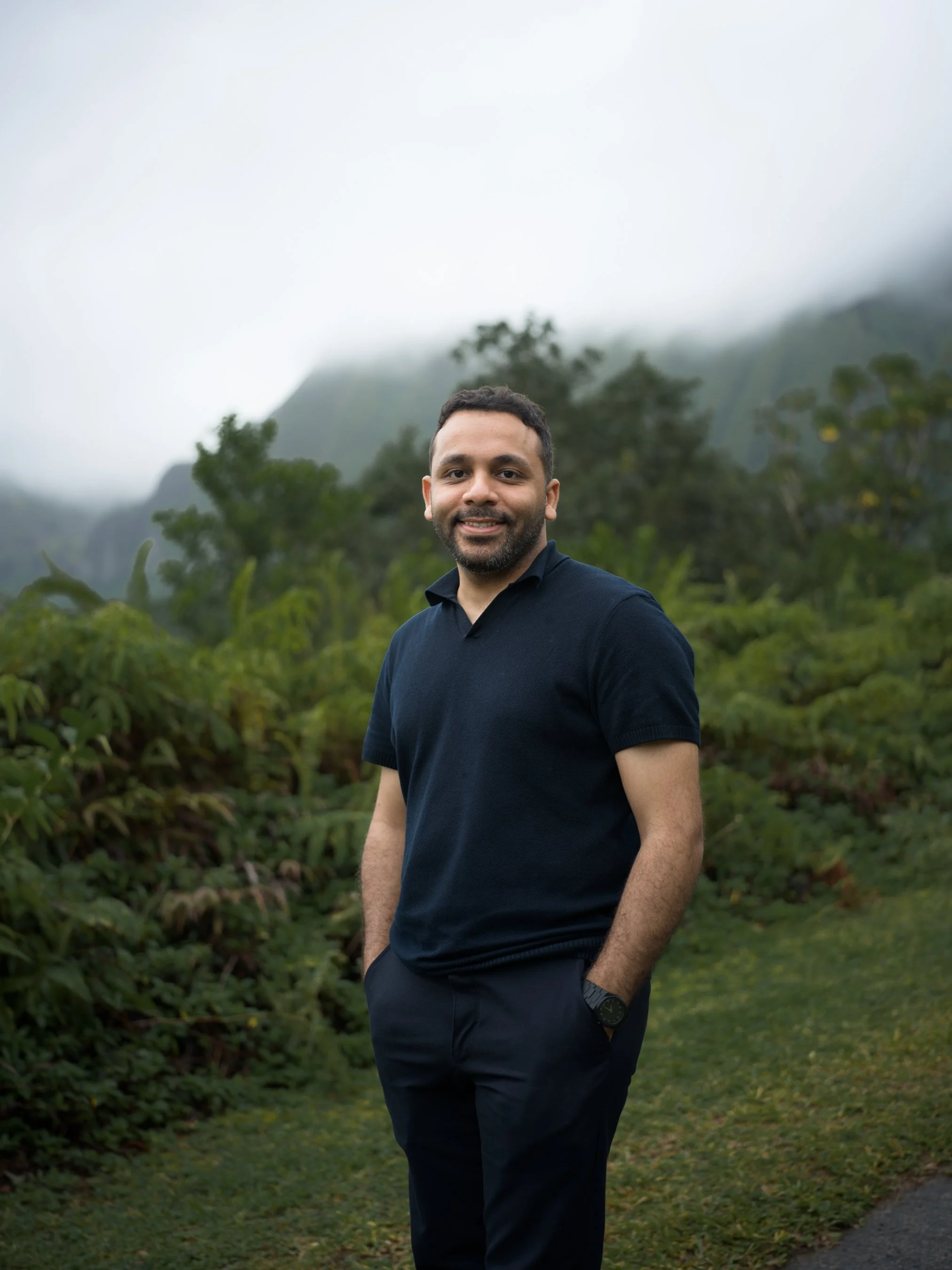 A man standing outdoors with a lush green background of trees and misty mountains, wearing a dark polo shirt and pants, smiling at the camera.