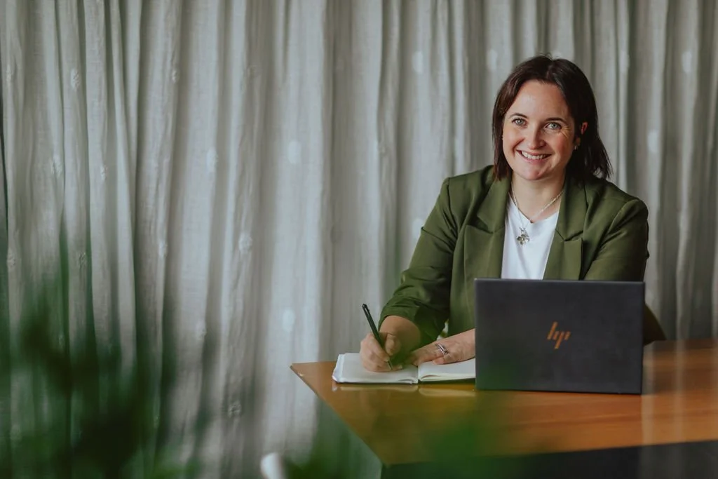 A woman sitting at a wooden table, smiling, with a laptop in front of her and a notebook she is writing in. Behind her is a curtain.