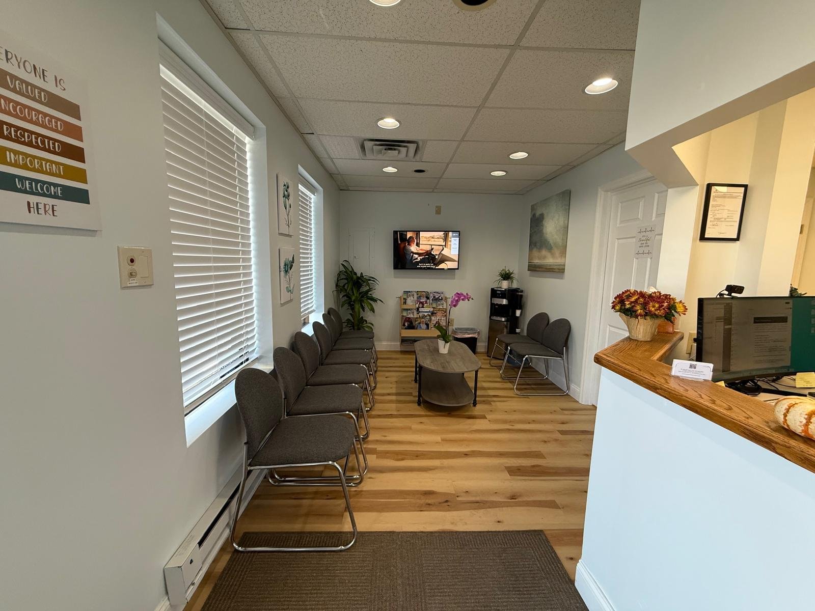 An empty waiting room with several gray chairs lined up against the walls, a small coffee table with a potted orchid, a colorful flower arrangement on the reception counter, a TV mounted on the wall, and light-colored wooden flooring.