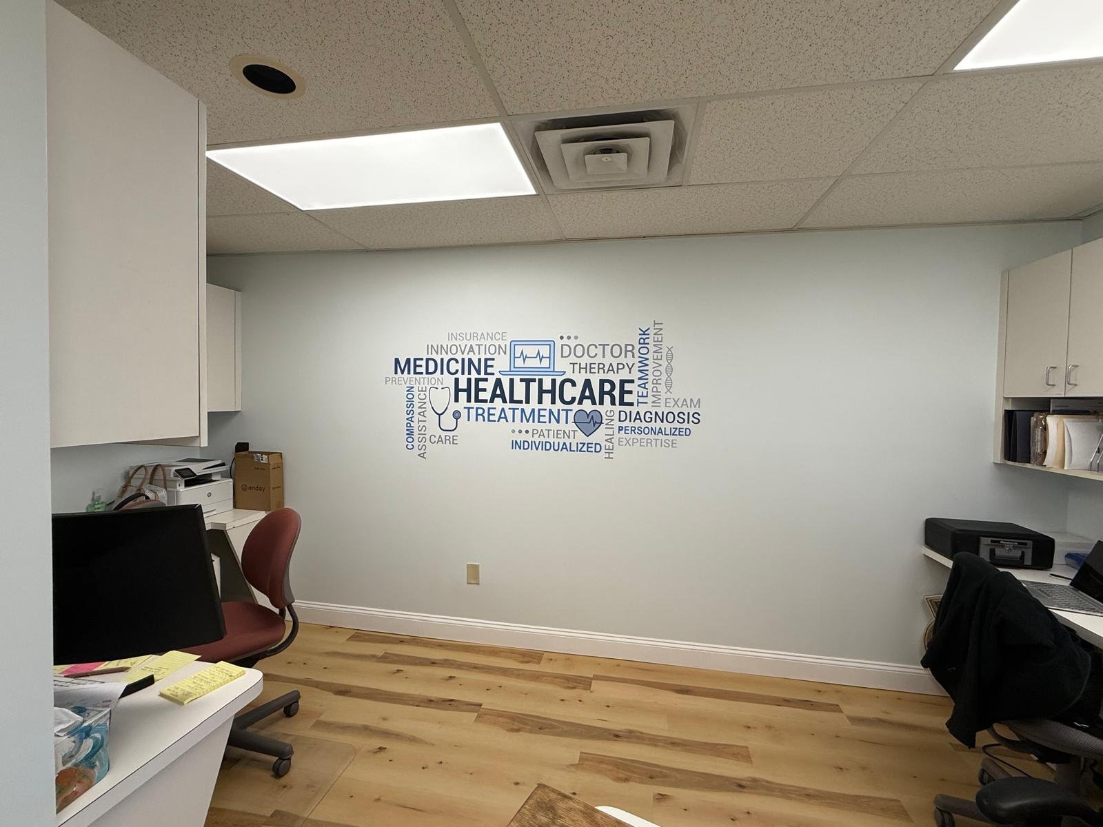 Office room with a wall decorated with healthcare-themed word cloud including 'Healthcare,' 'Medicine,' 'Diagnosis,' and 'Treatment' with desks, chairs, and office supplies.