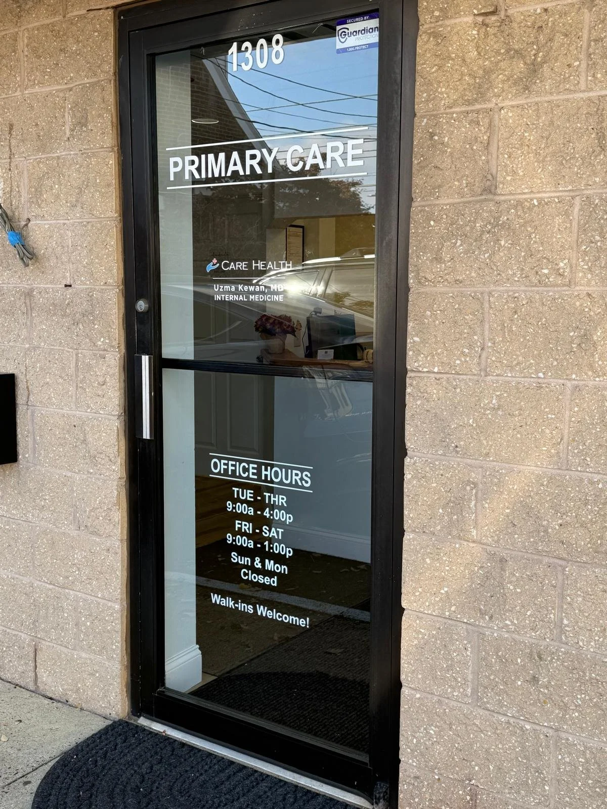 Glass door with office hours and primary care information for a healthcare clinic, labeled 1308, with brick wall exterior, and reflections of parked cars and power lines.