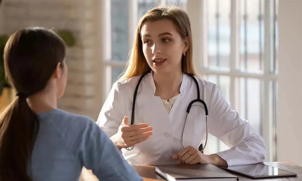 A young female doctor with a stethoscope discusses with a female patient in a medical office with large windows.