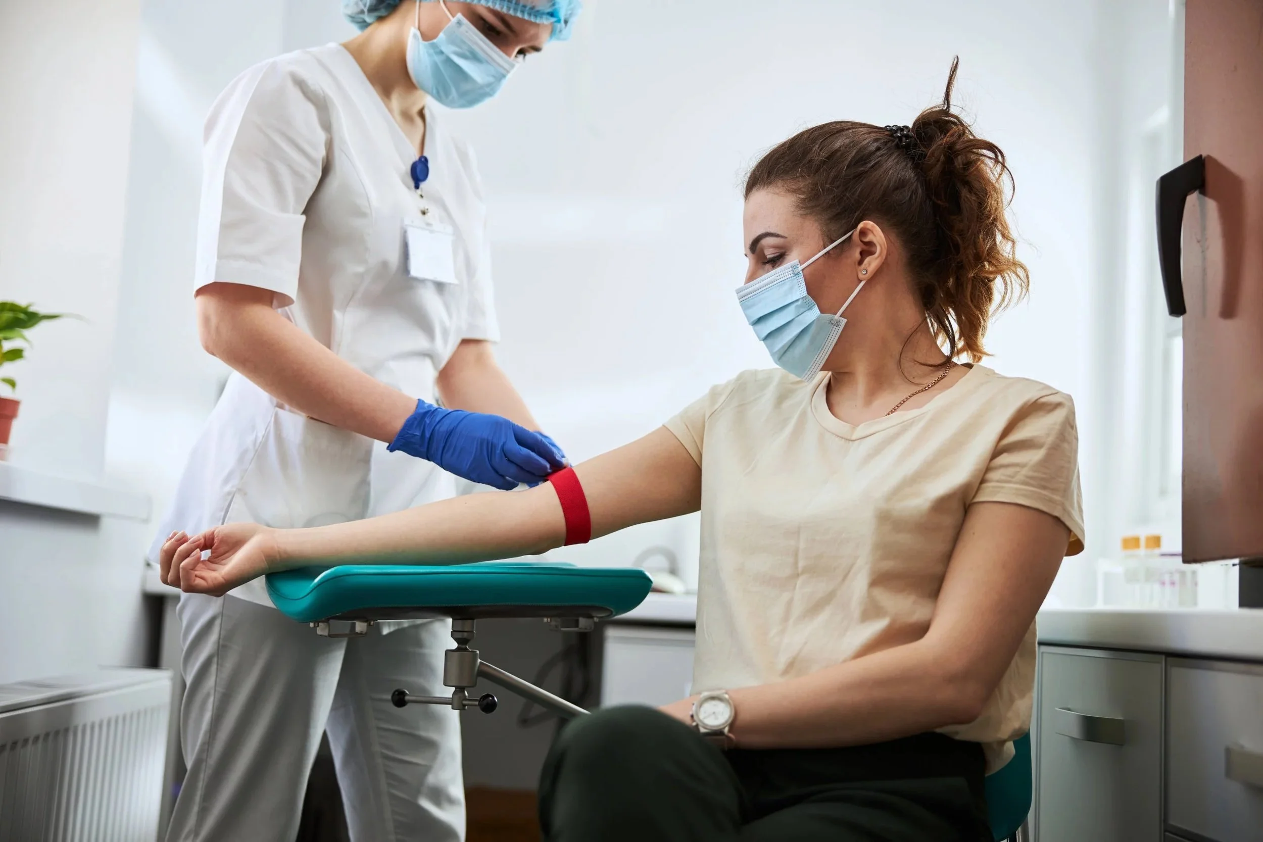 A woman with a face mask and beige shirt is donating blood in a medical clinic, with a healthcare worker in scrubs and face mask preparing her arm.