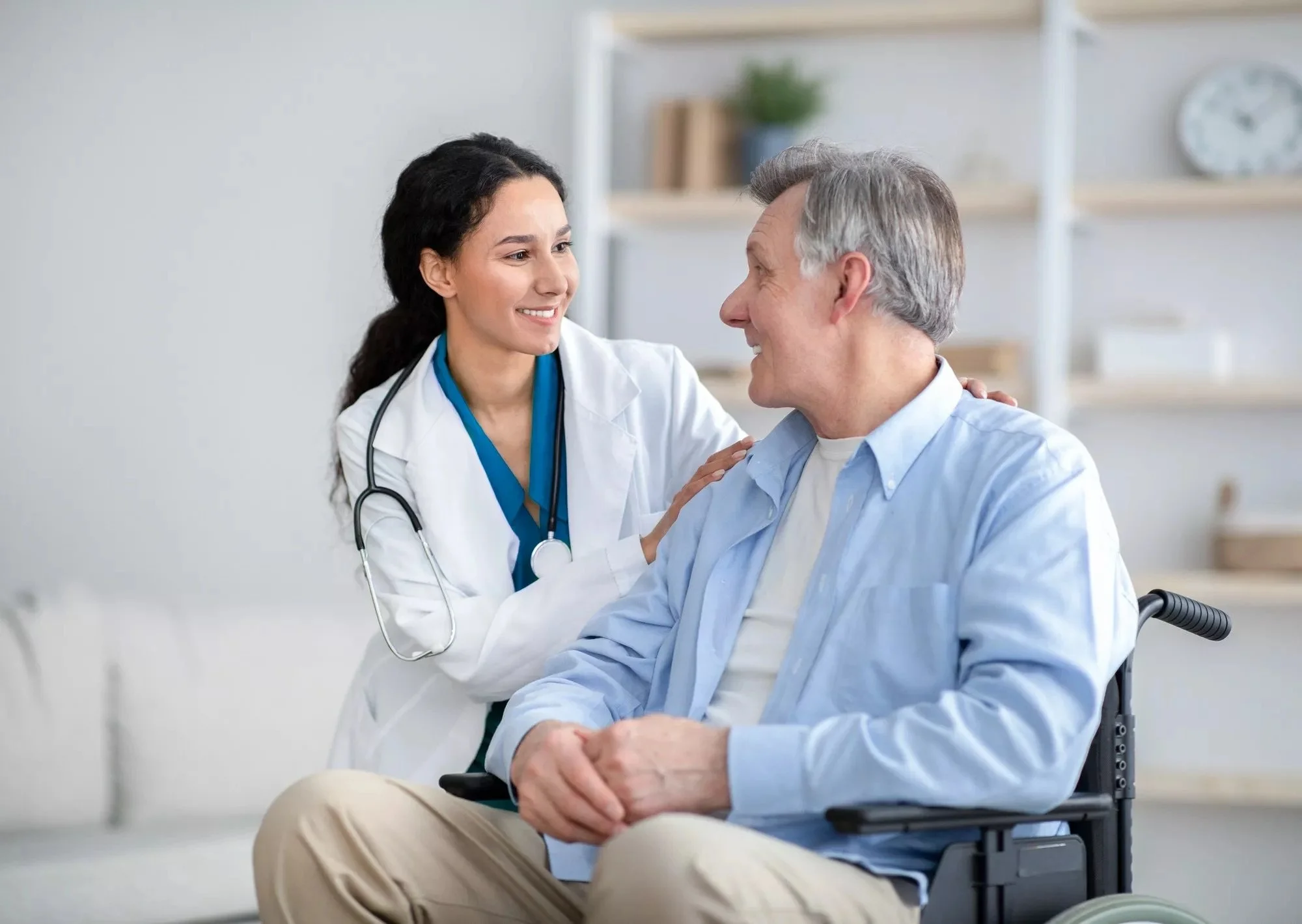 A female healthcare professional in a white coat talking to an elderly male patient in a wheelchair in a medical office.