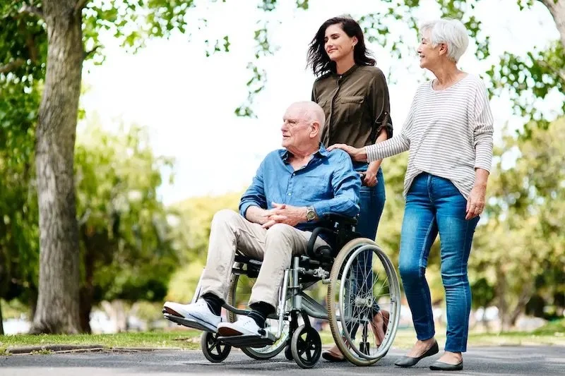 Three women, one in a wheelchair, walking outdoors among trees on a bright day.