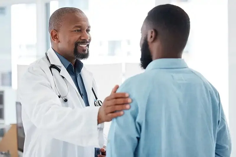 A doctor in a white coat with a stethoscope on his neck smiling and placing his hand on a patient's shoulder during a consultation in a medical office with large windows.