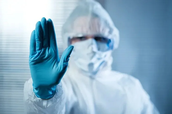 Medical worker in protective gear with a face mask, goggles, and blue gloves, raising a hand as a stop gesture.