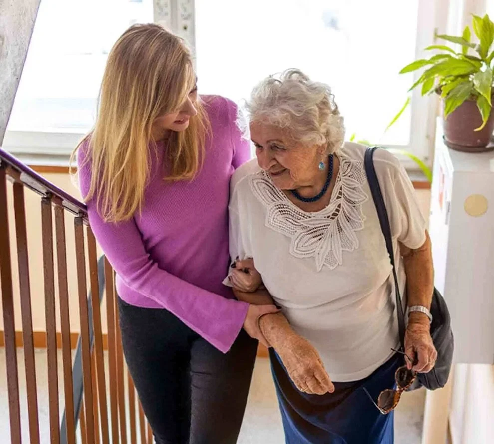 A young woman helping an elderly woman walk, holding her arm inside a home with natural light, a plant on a white cabinet nearby.