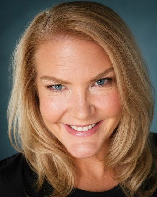Close-up of a smiling woman with blonde hair and blue eyes against a teal background.