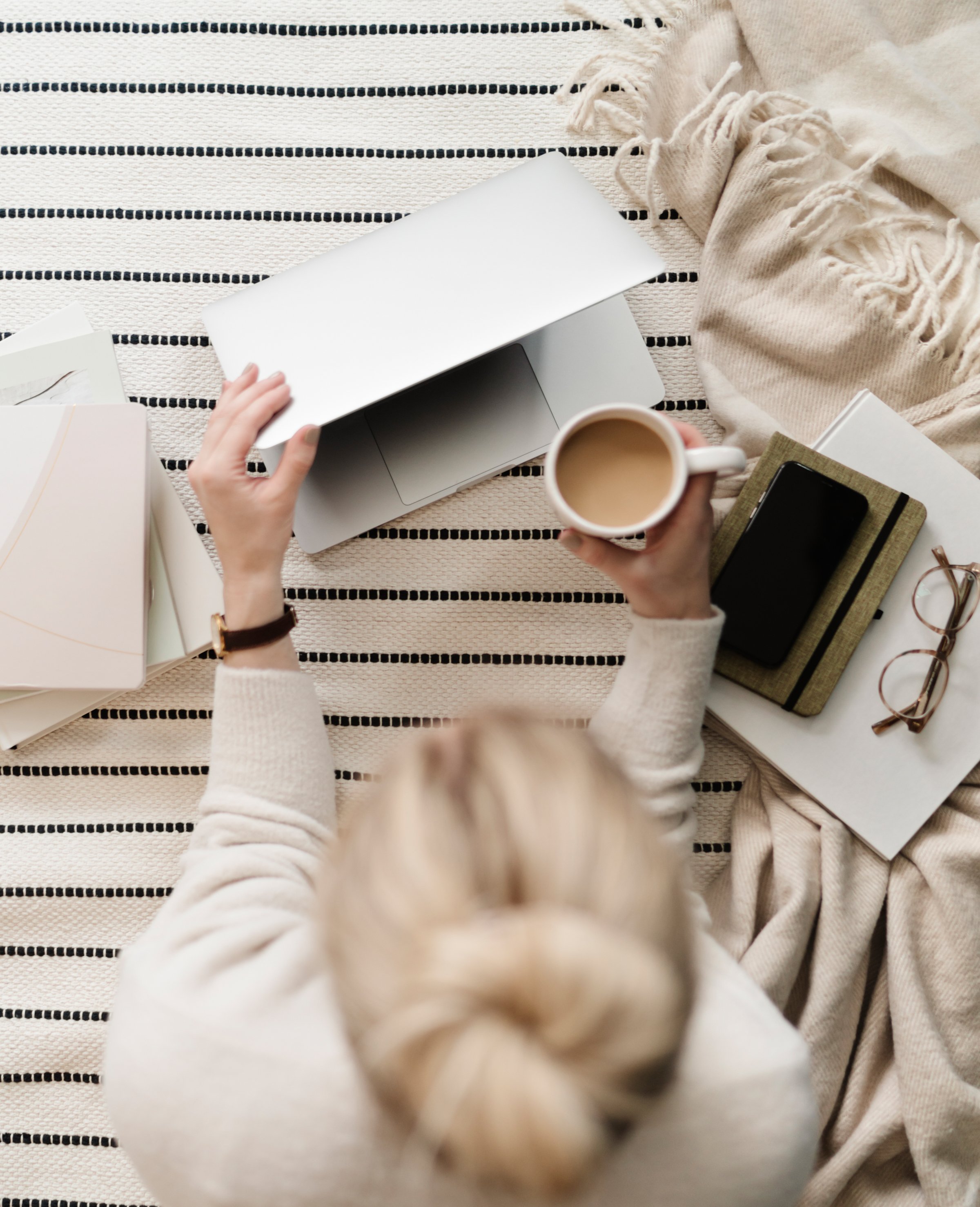 Overhead view of a woman sitting on a striped rug with a laptop, a cup of coffee, glasses, a smartphone, notebooks, and papers around her.