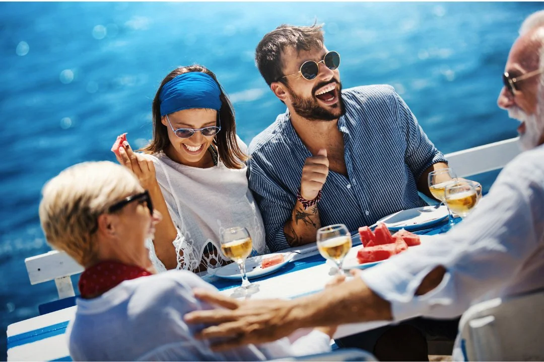 Four people enjoying a meal together on a boat with the ocean in the background, laughing and smiling, with glasses of white wine and slices of watermelon on the table.