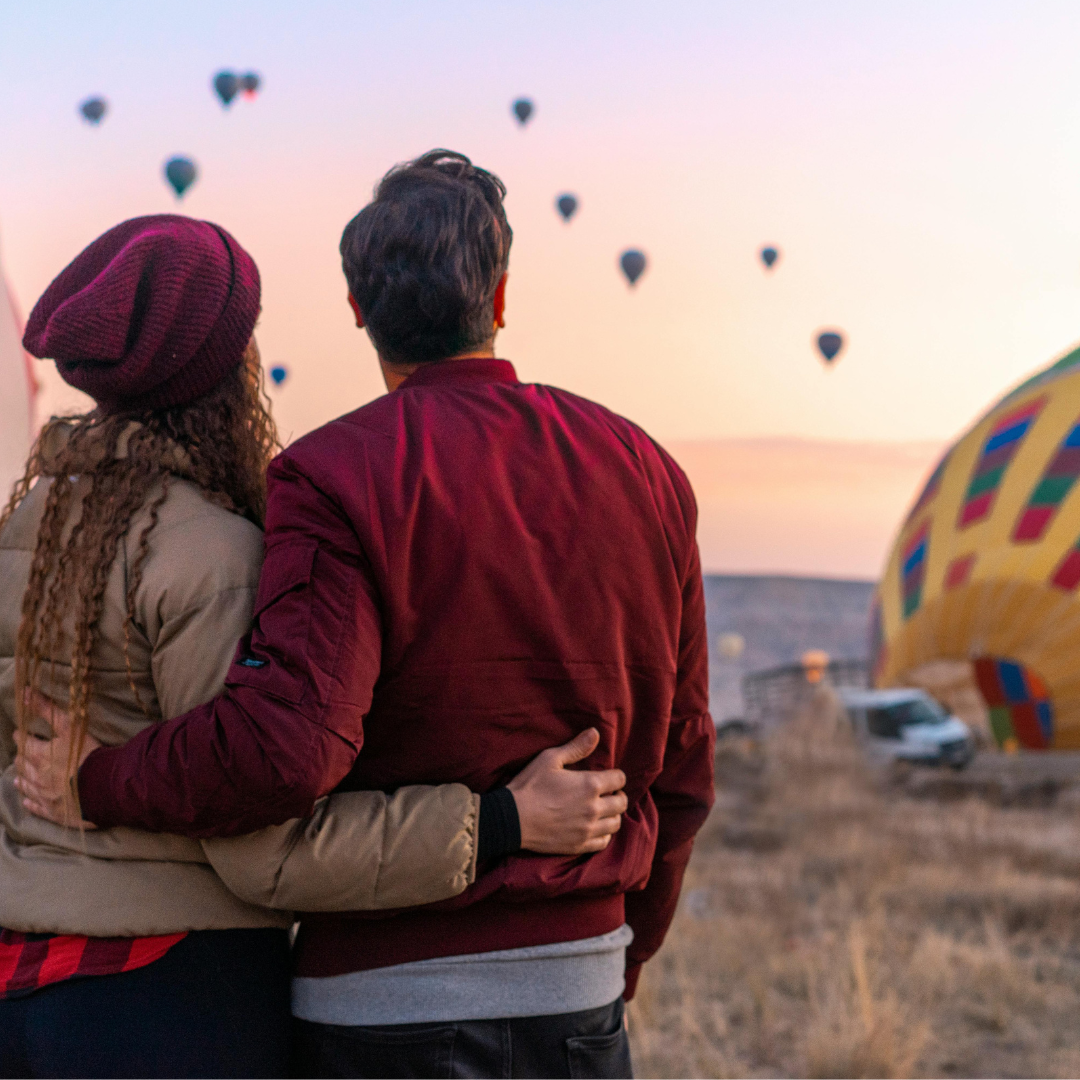 A couple standing close together, watching hot air balloons ascend into the sky at sunset or sunrise in an open field.