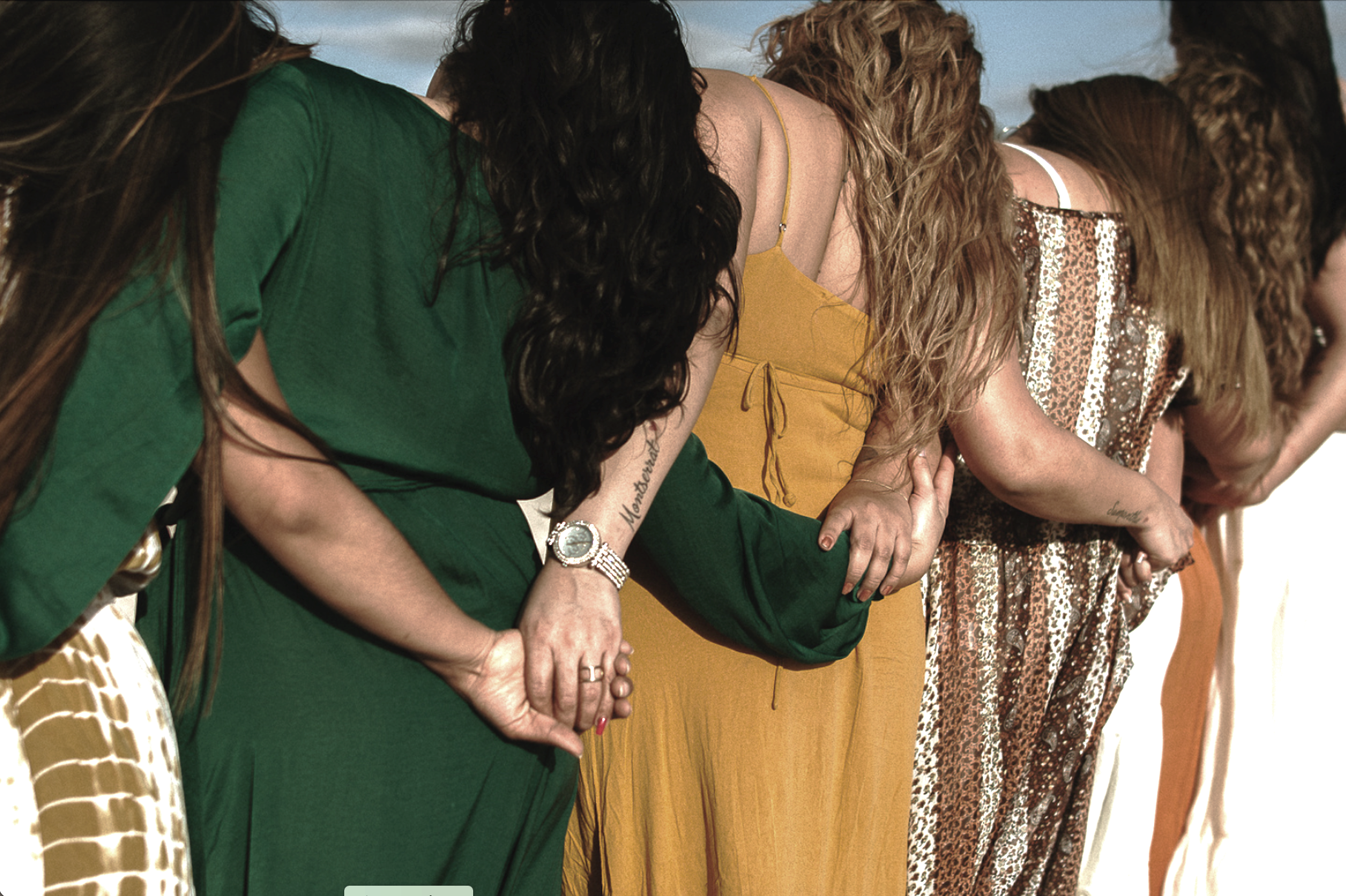 Six women holding hands in a line, dressed in colorful outfits, some with curly or straight hair.