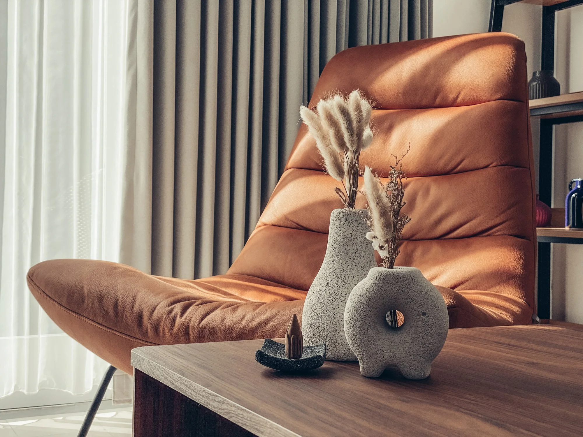 Living room with a tan leather chair, a textured white vase with dried flowers, and another textured vase, on a wooden table near a window with sheer curtains and black drapes.
