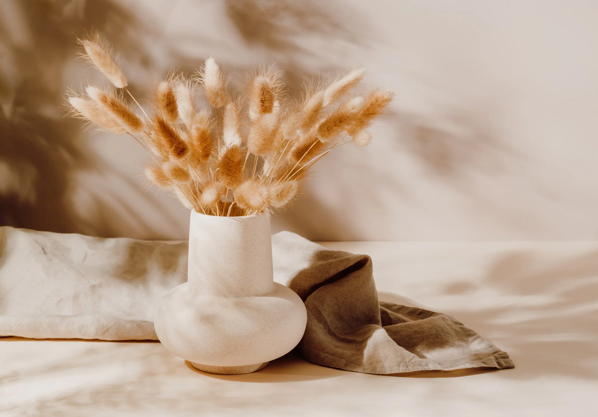 A white vase with beige dried bunny tail grass arranged inside, placed on a neutral fabric surface with soft lighting.