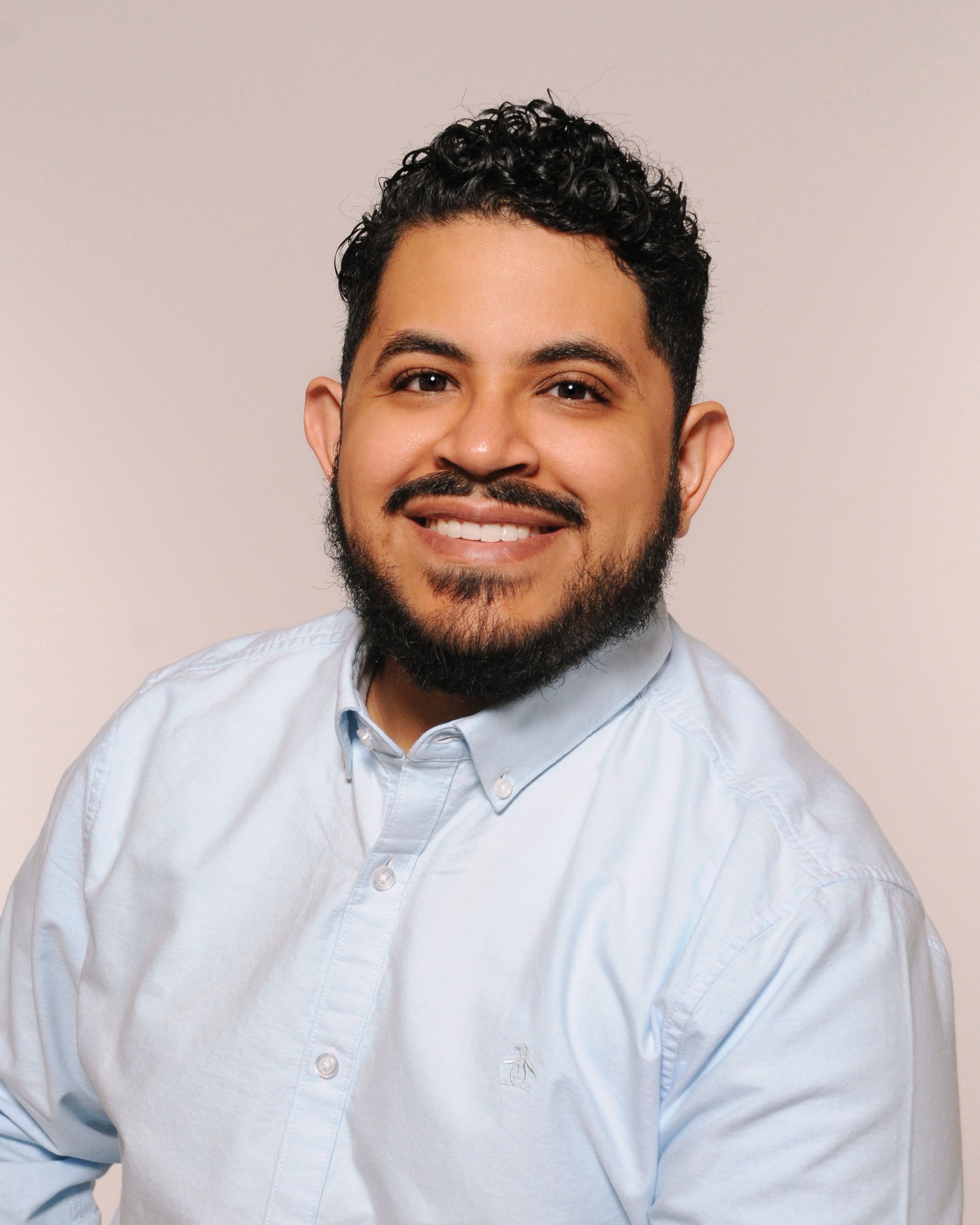 Close-up of a smiling man with dark curly hair and facial hair, wearing a light blue button-up shirt against a plain, light-colored background.