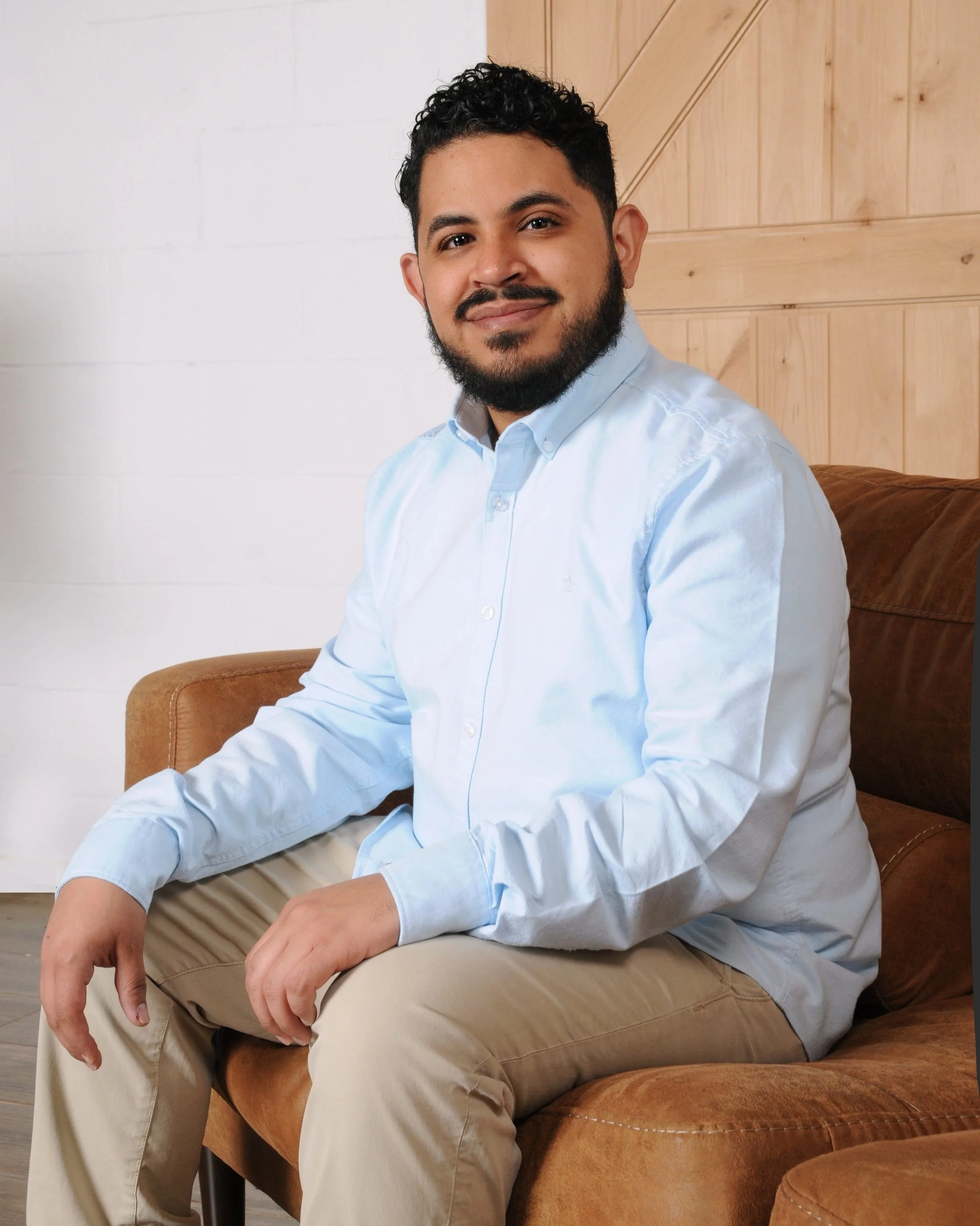 A man sitting on a brown sofa, wearing a light blue shirt and khaki pants, smiling at the camera with a background of a wooden wall and white wall.