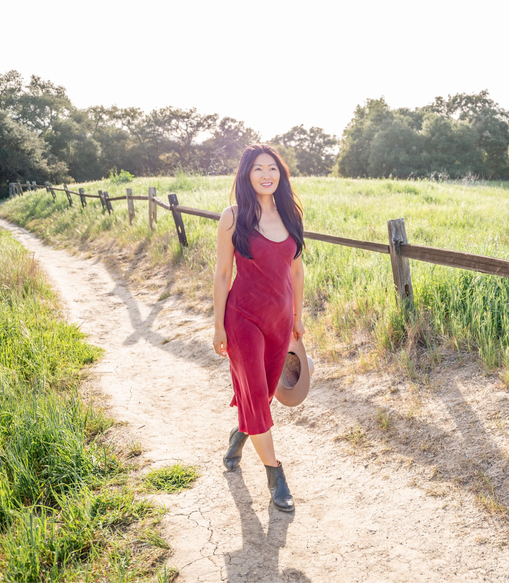 Woman walking along a dirt path through a green field with trees in the background, carrying a hat.