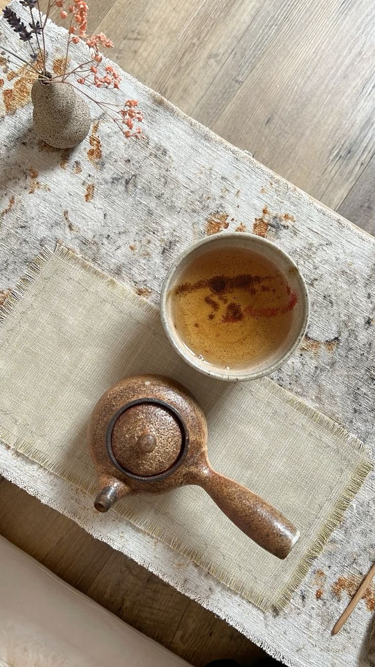 A textured ceramic teapot, a cup of tea or broth, and a small vase with dried pink and purple flowers on a distressed white table with a beige cloth placemat.