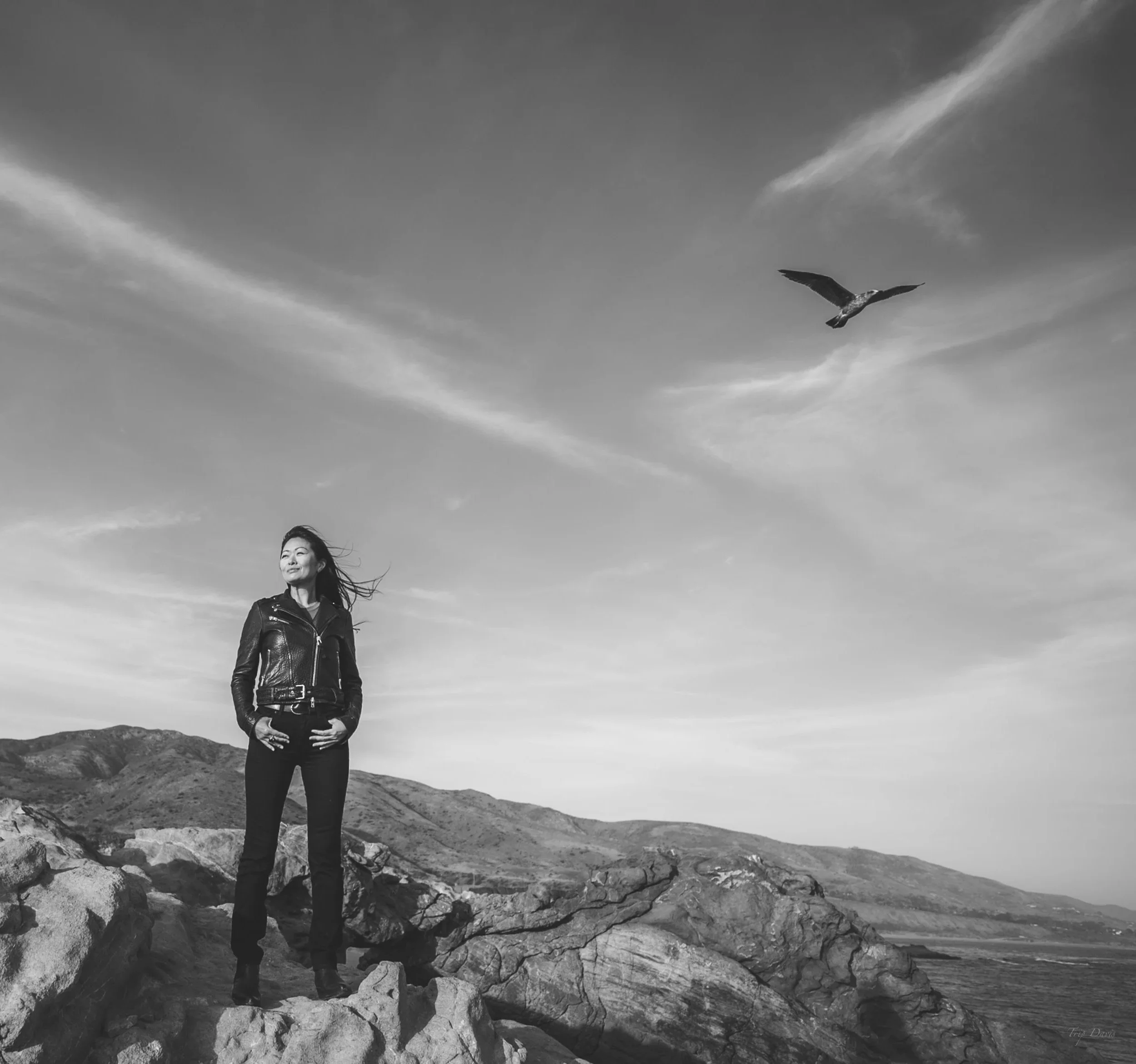 A psychic, intuitive woman standing on rocks by the water with mountains in the background, relaxed and smiling, with a cloudy sky and a bird flying overhead. Mother Earth, cosmos, heart coherence.