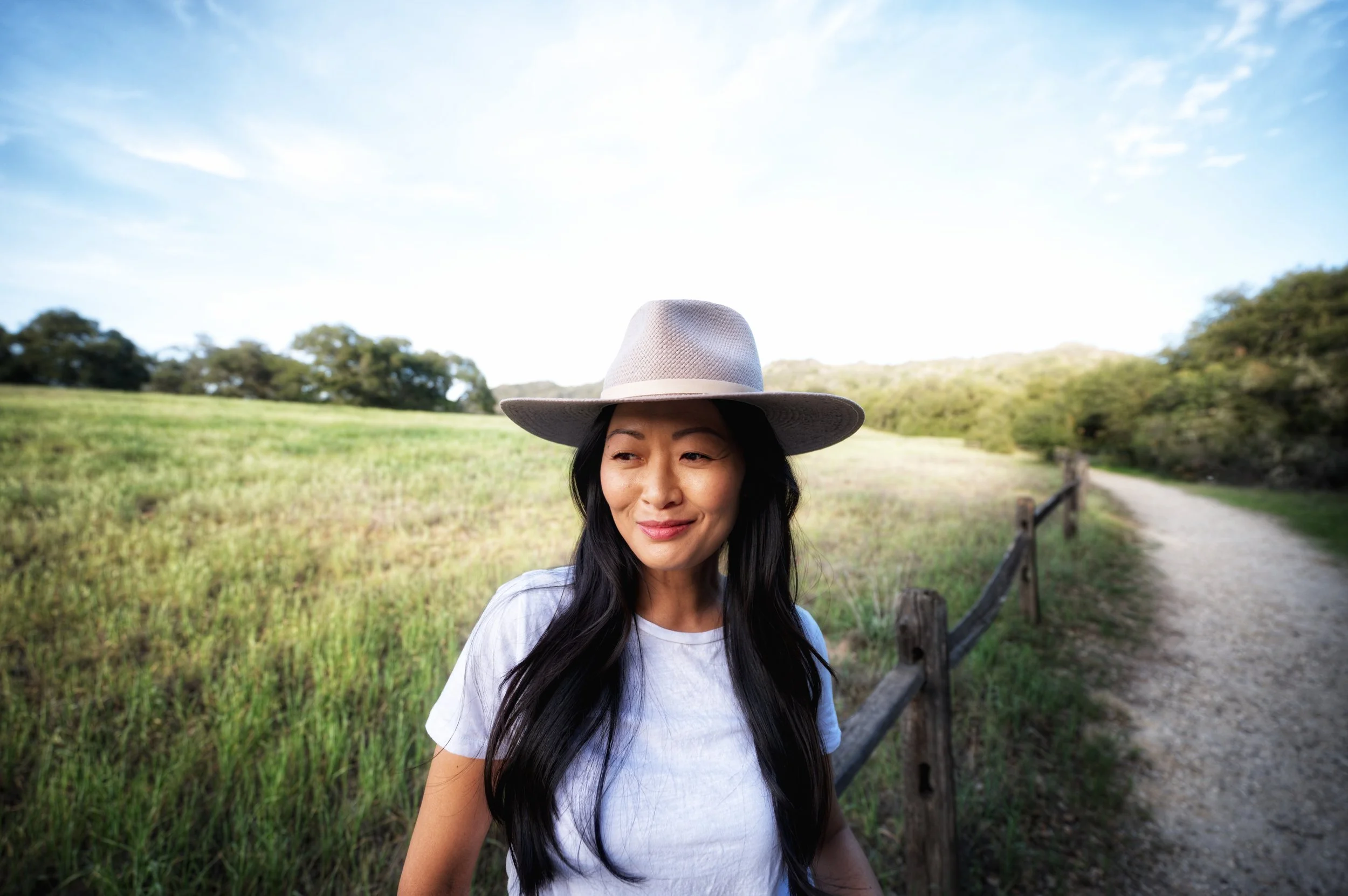 A woman with long dark hair wearing a white hat and white T-shirt, standing outdoors on a pathway with a grassy field and trees in the background, under a partly cloudy sky.