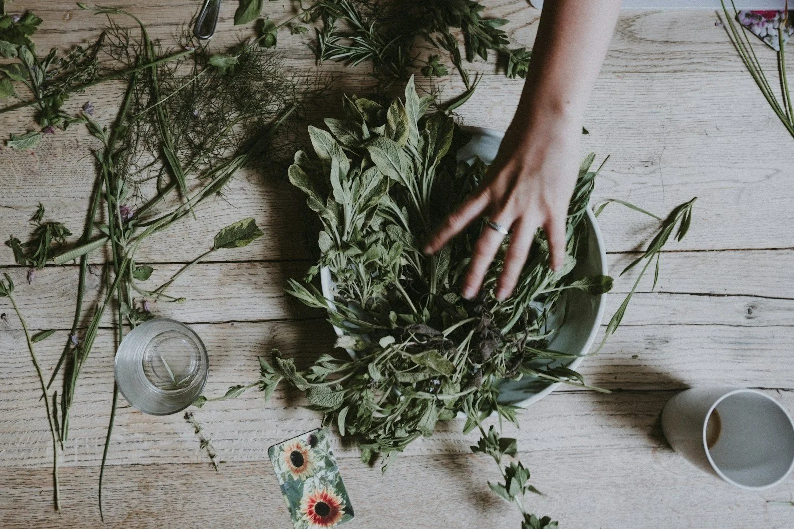 Person arranging various fresh herbs on a wooden table, with a small card featuring sunflowers, a glass jar, and a white cup nearby.