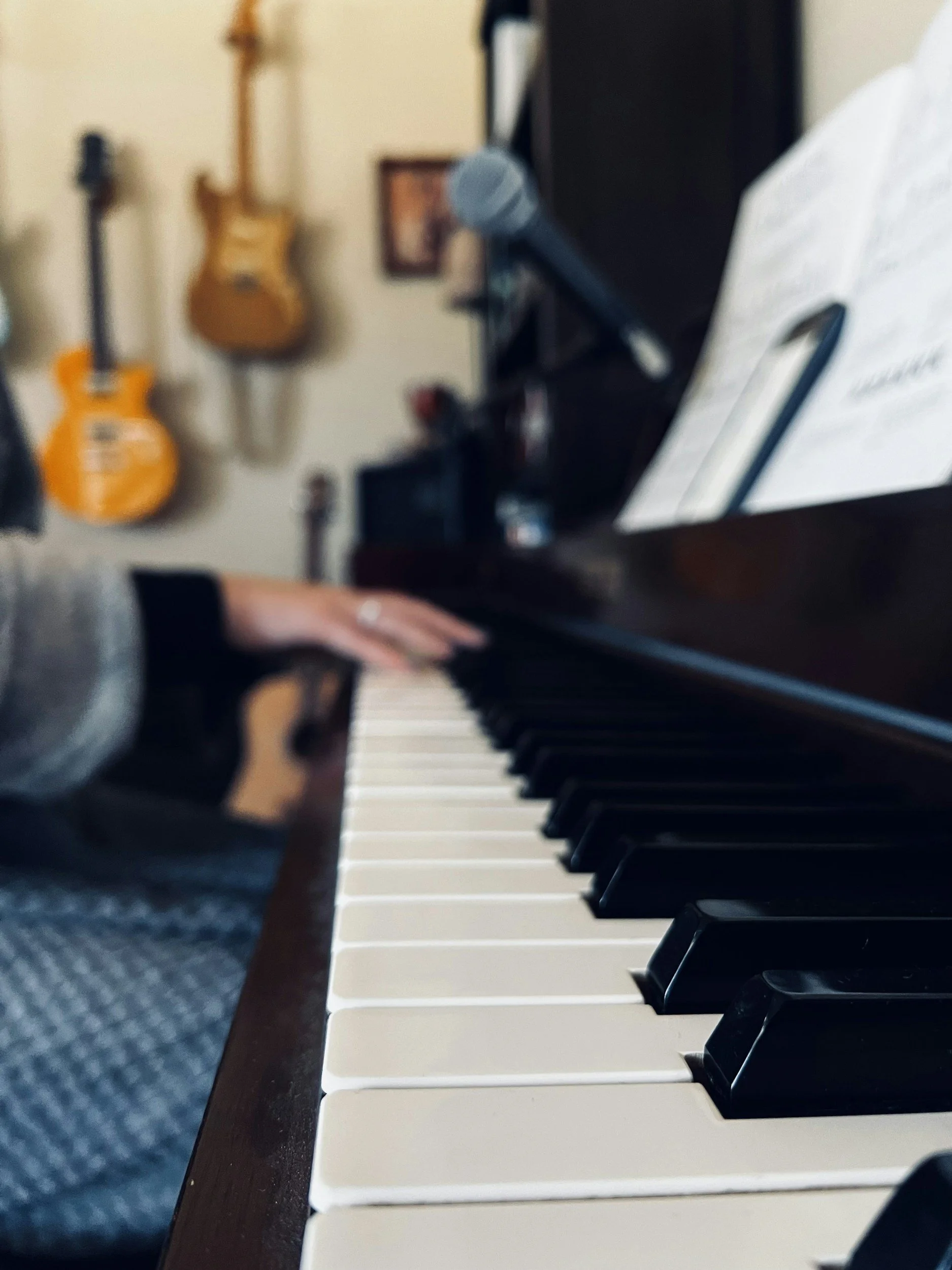 Close-up of a piano keyboard with a person's hand pressing the keys, in a room with guitars hanging on the wall and a microphone on a stand.