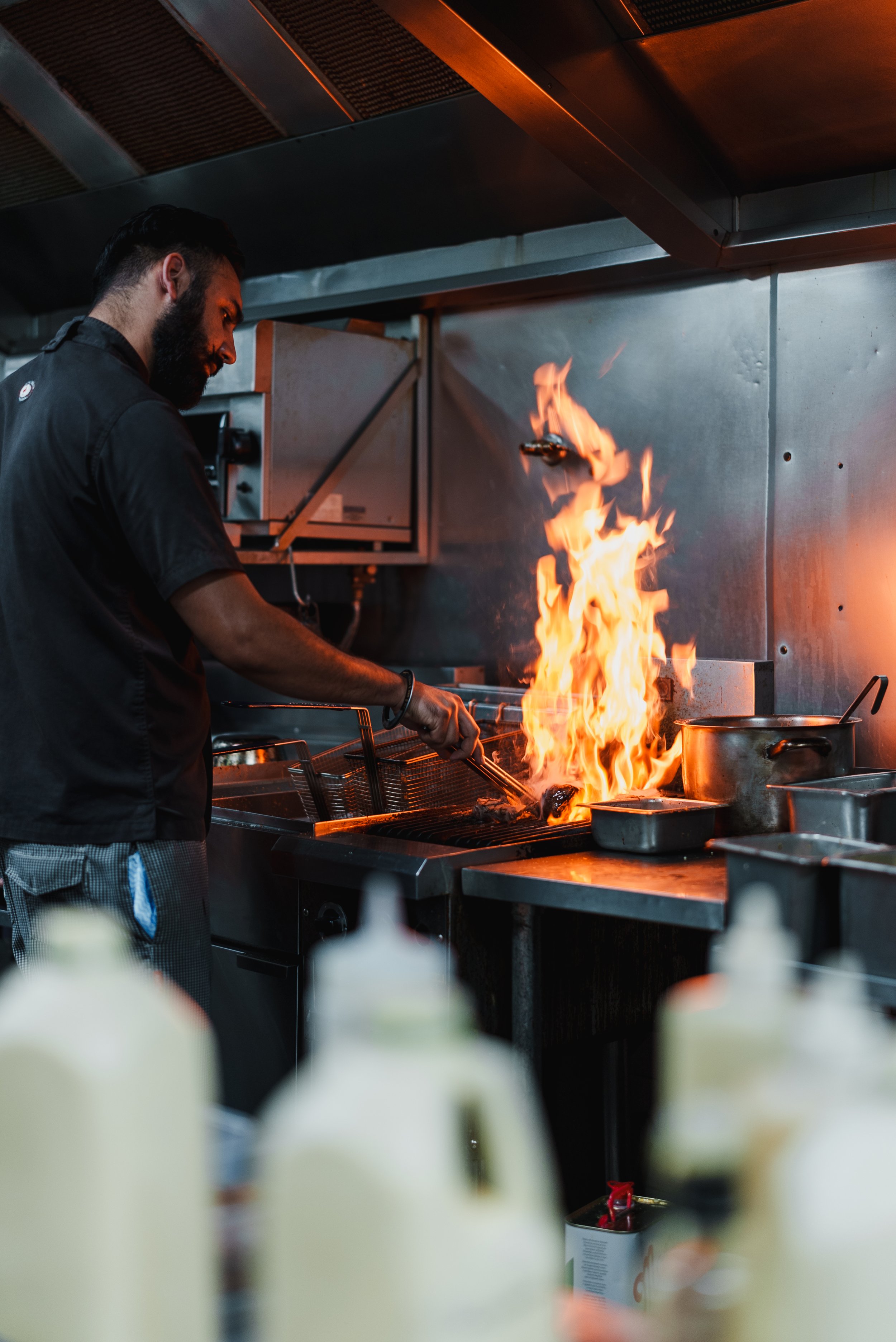 Chef cooking with high flames in a professional kitchen, using tongs to handle food on the stove.